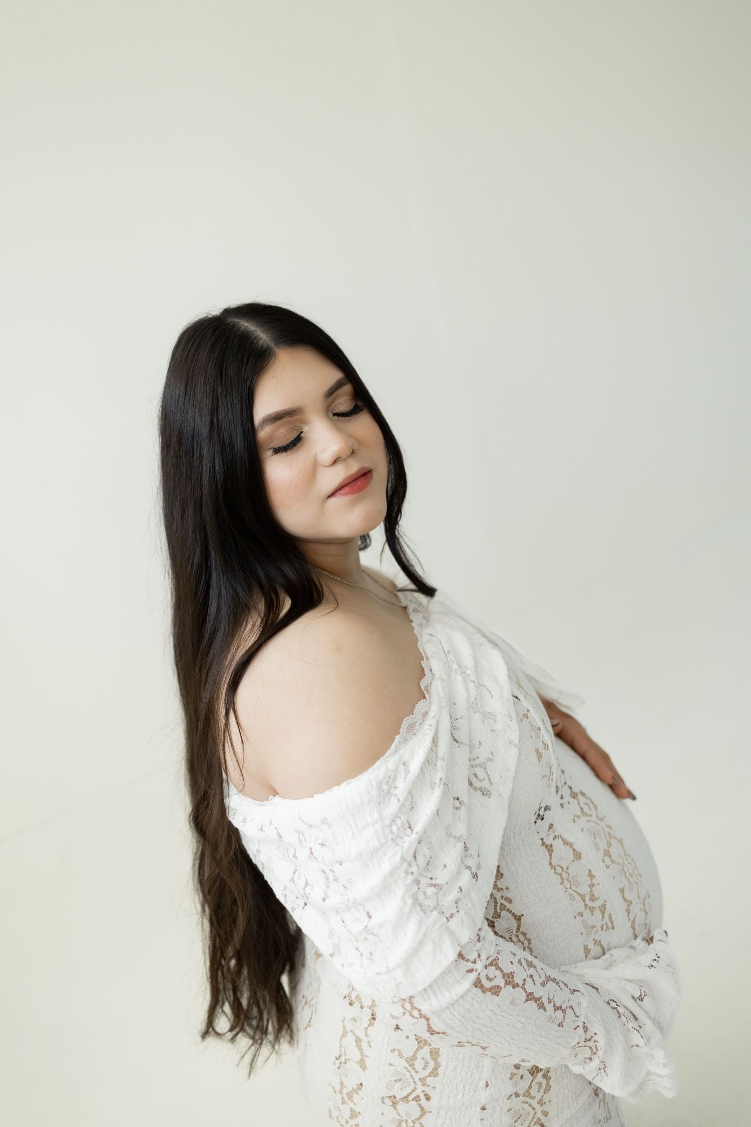 Elegant portrait against light background showing long dark hair and delicate white lace dress with off-shoulder design.