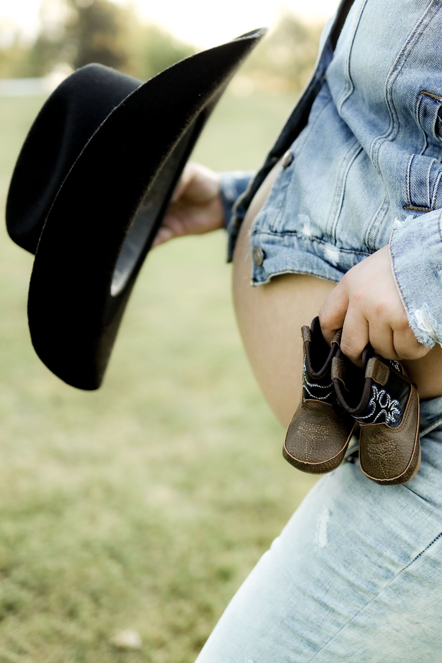 A denim-clad figure holds tiny brown boots and a black cowboy hat against a soft outdoor background.
