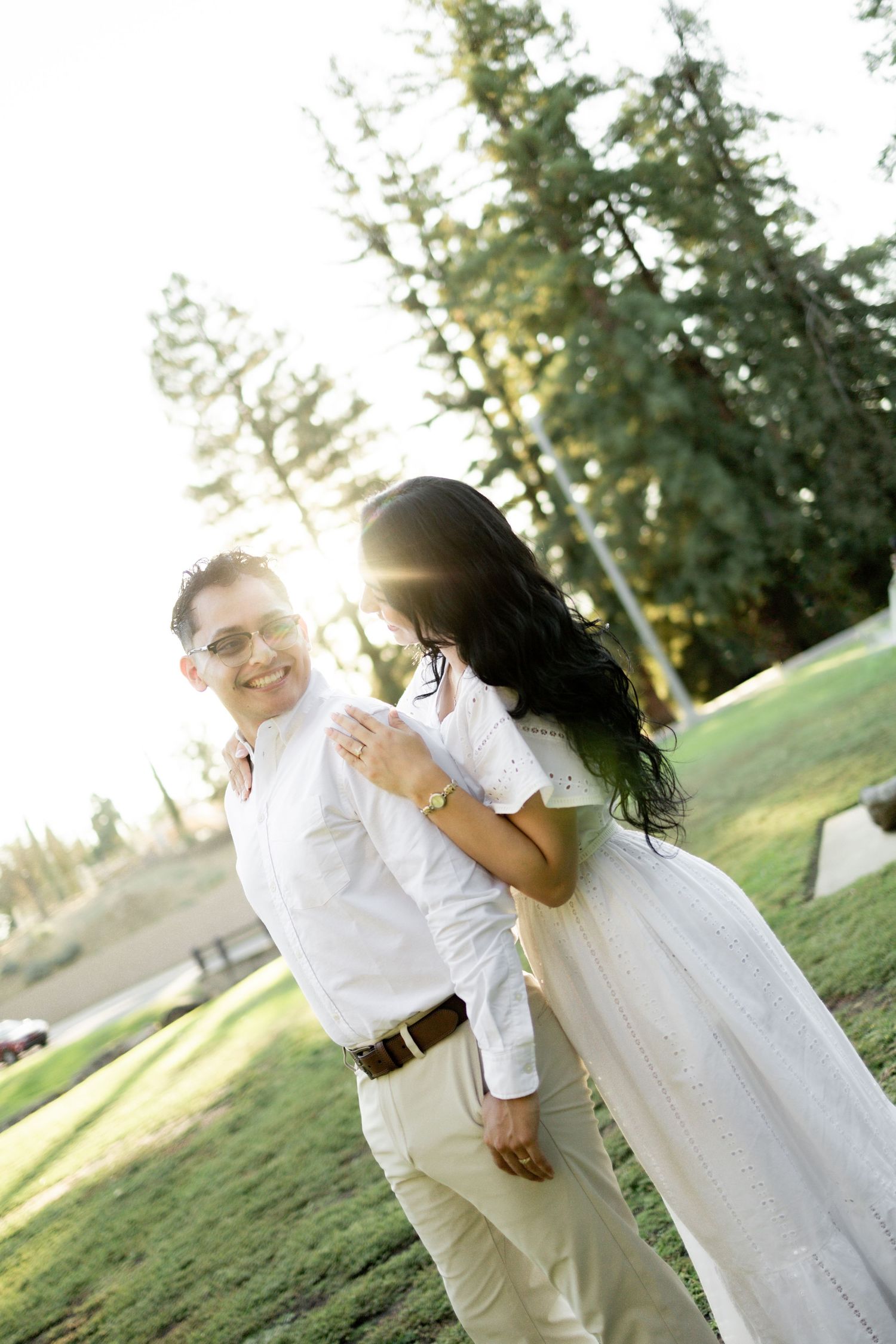 A joyful couple laughing together in white outfits against sunlit trees.