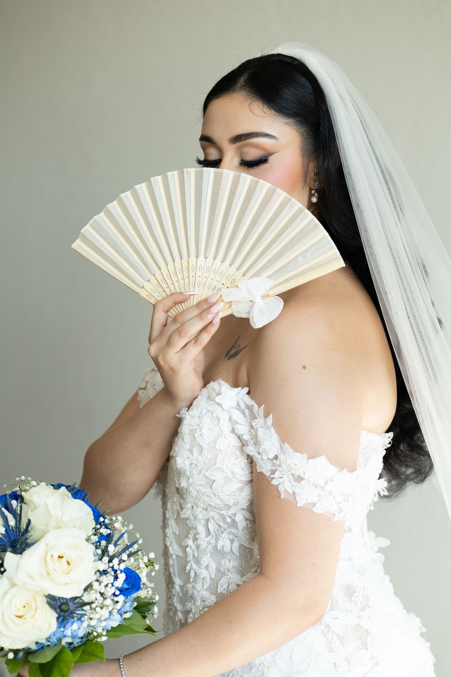 Elegant bride holding white decorative fan with lace dress detail and blue floral bouquet.