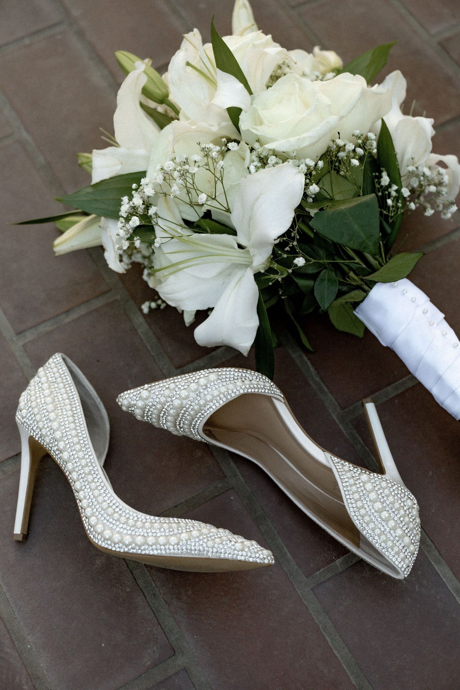 Elegant white bridal shoes with crystal embellishments displayed alongside white floral boutonnieres on dark surface.