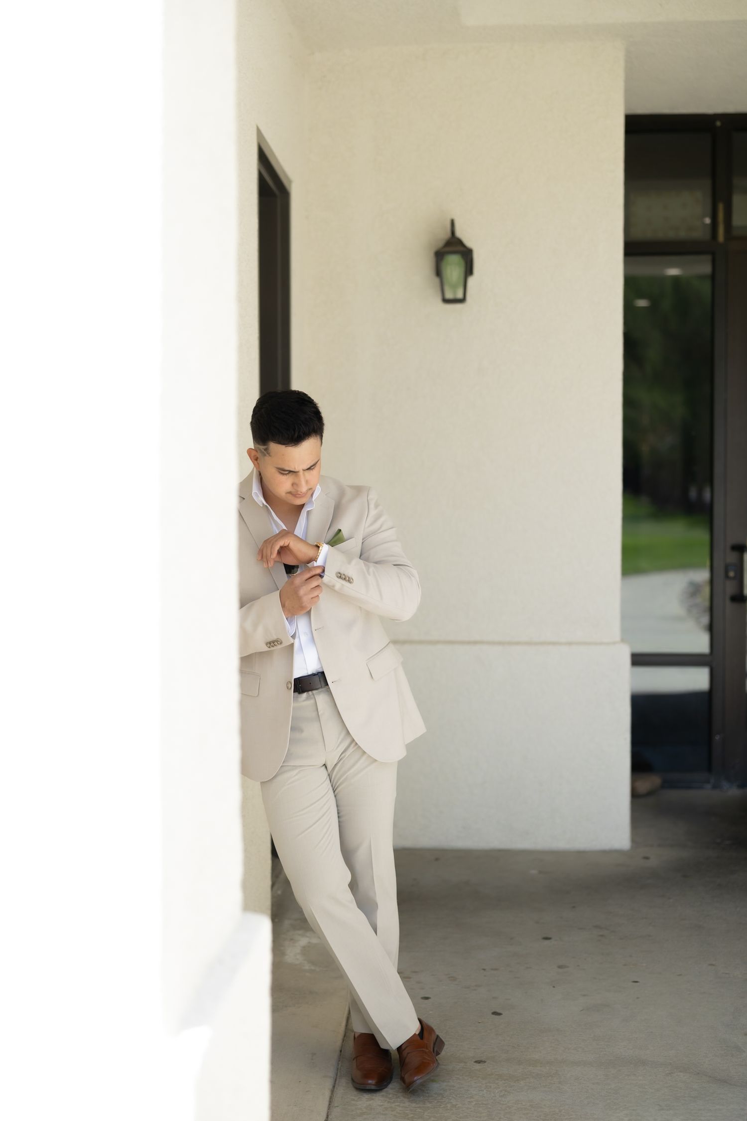 A well-dressed man in a beige suit leans against a white pillar, looking down at his phone outdoors.