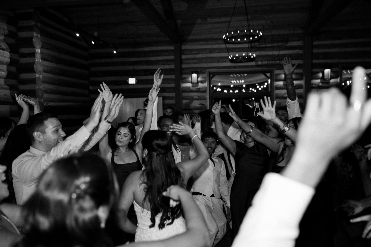 Black and white photo of wedding guests dancing with hands raised in a rustic log cabin venue.
