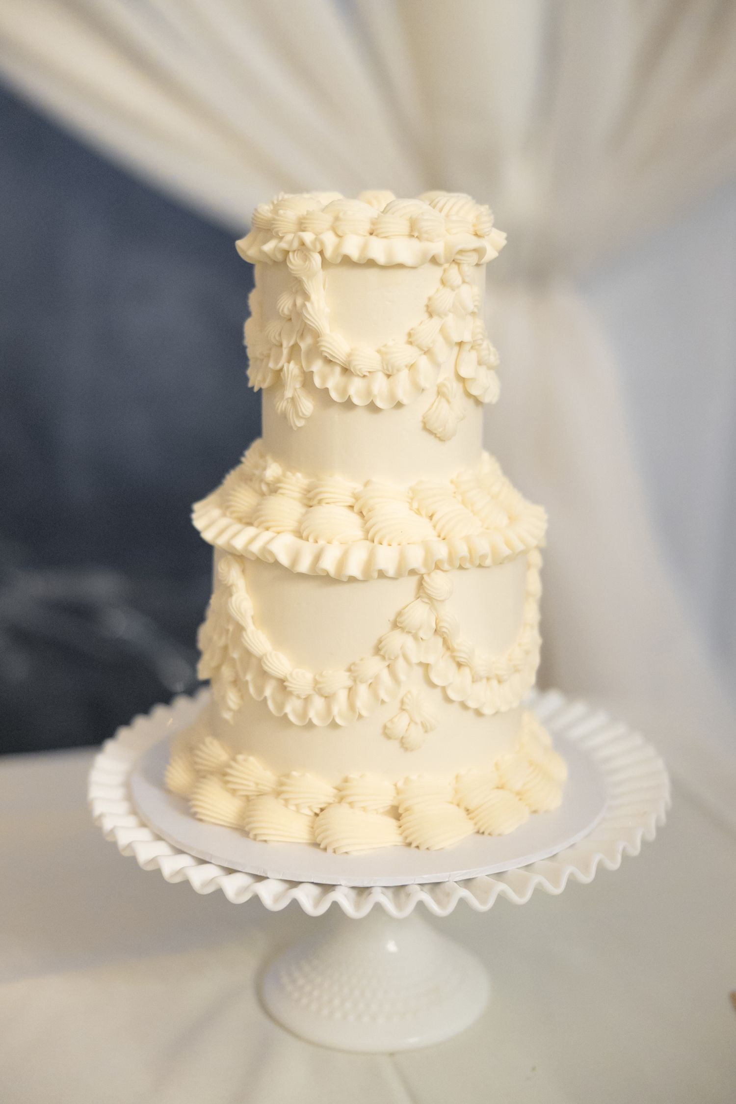 Elegant three-tier white wedding cake with ruffled buttercream details on milk glass stand.