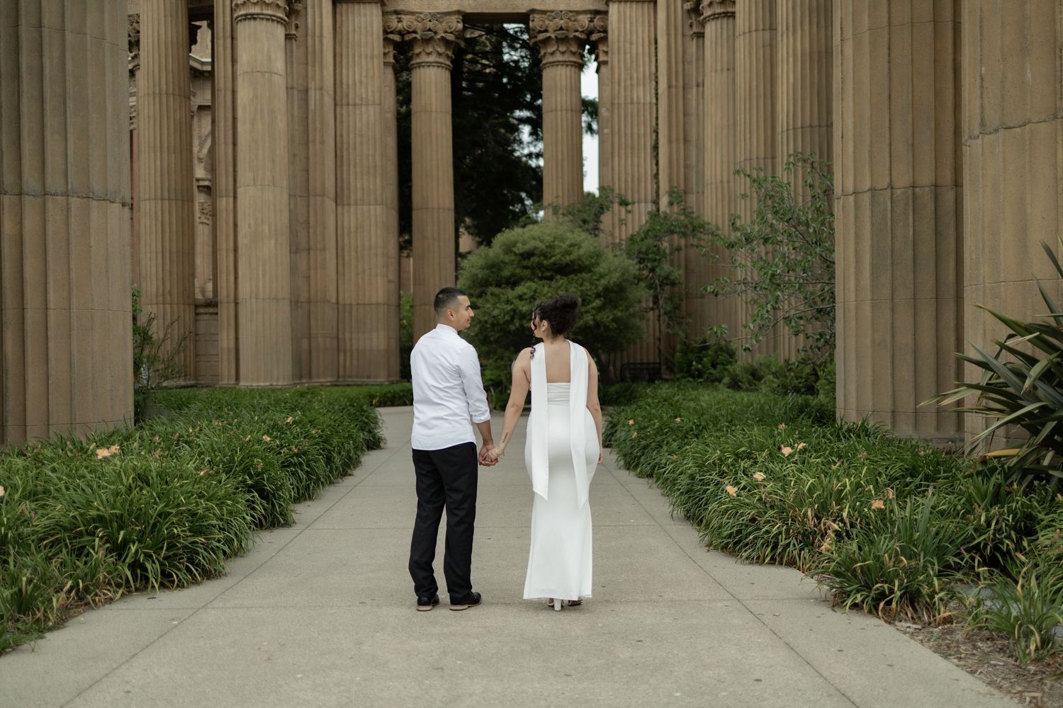 A couple stands between stone columns in a garden setting during a wedding ceremony.