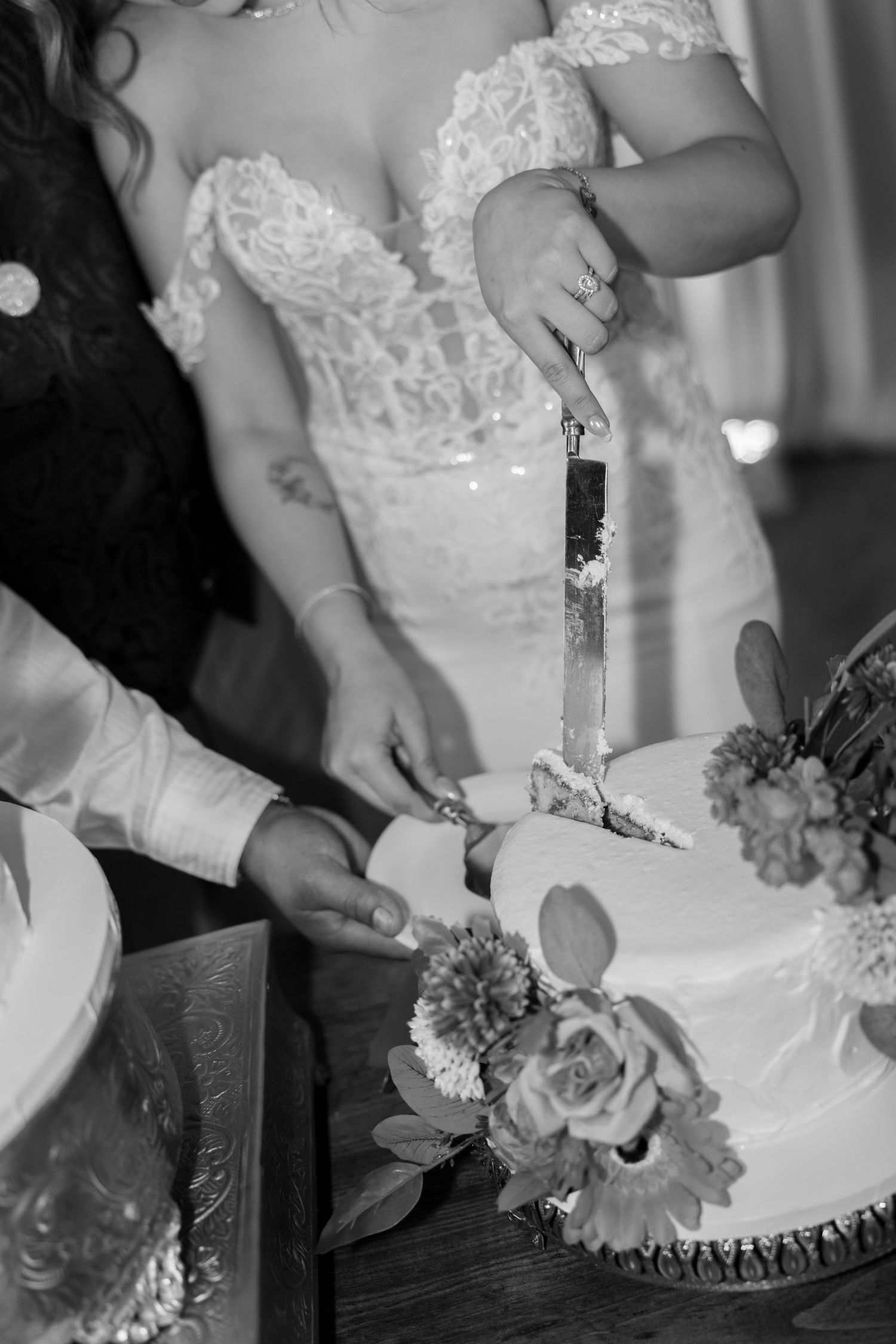 Black and white photo of bride and groom cutting their floral-decorated wedding cake together.