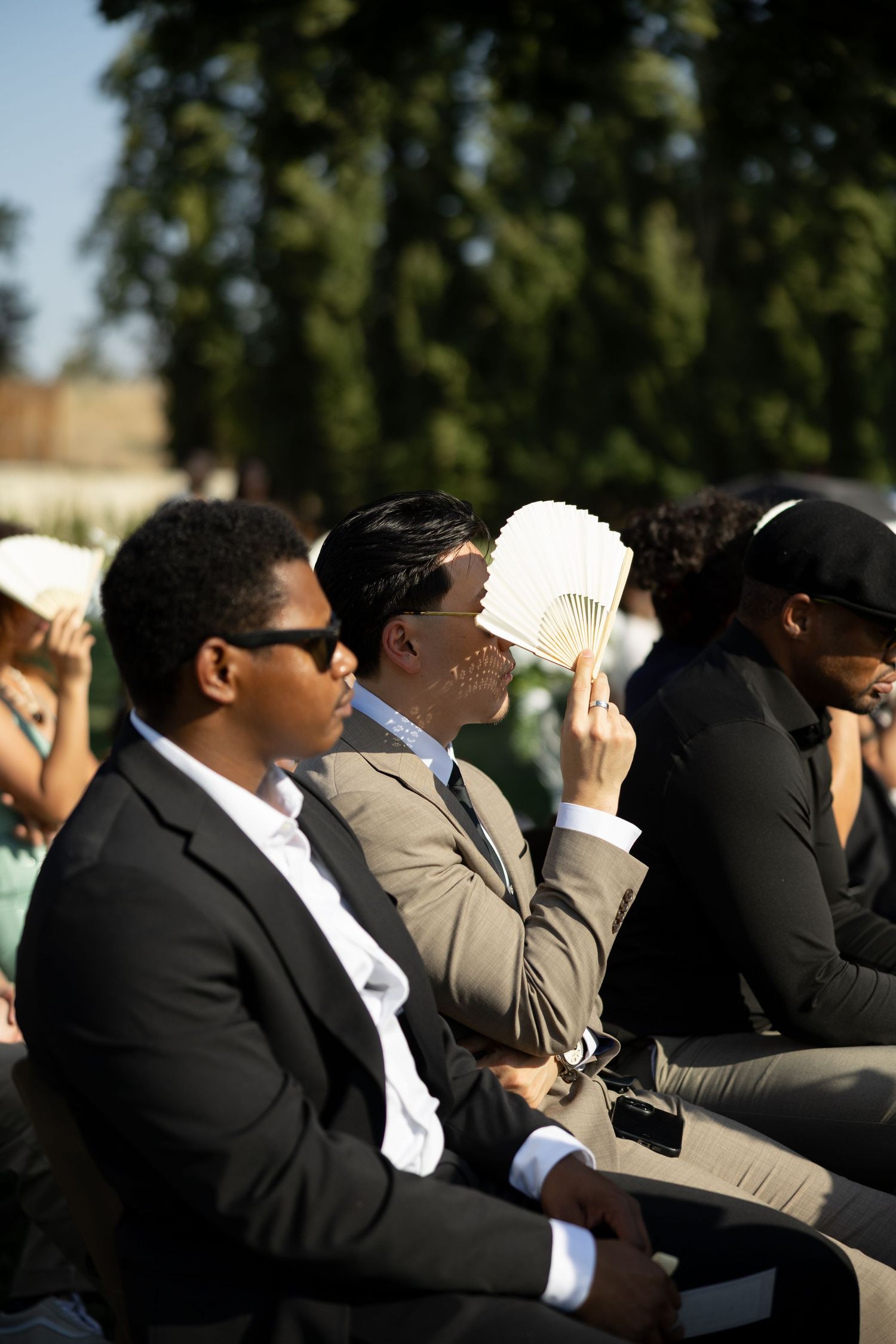 Wedding guests in suits sit outdoors in sunlight, one fanning himself during an outdoor ceremony.