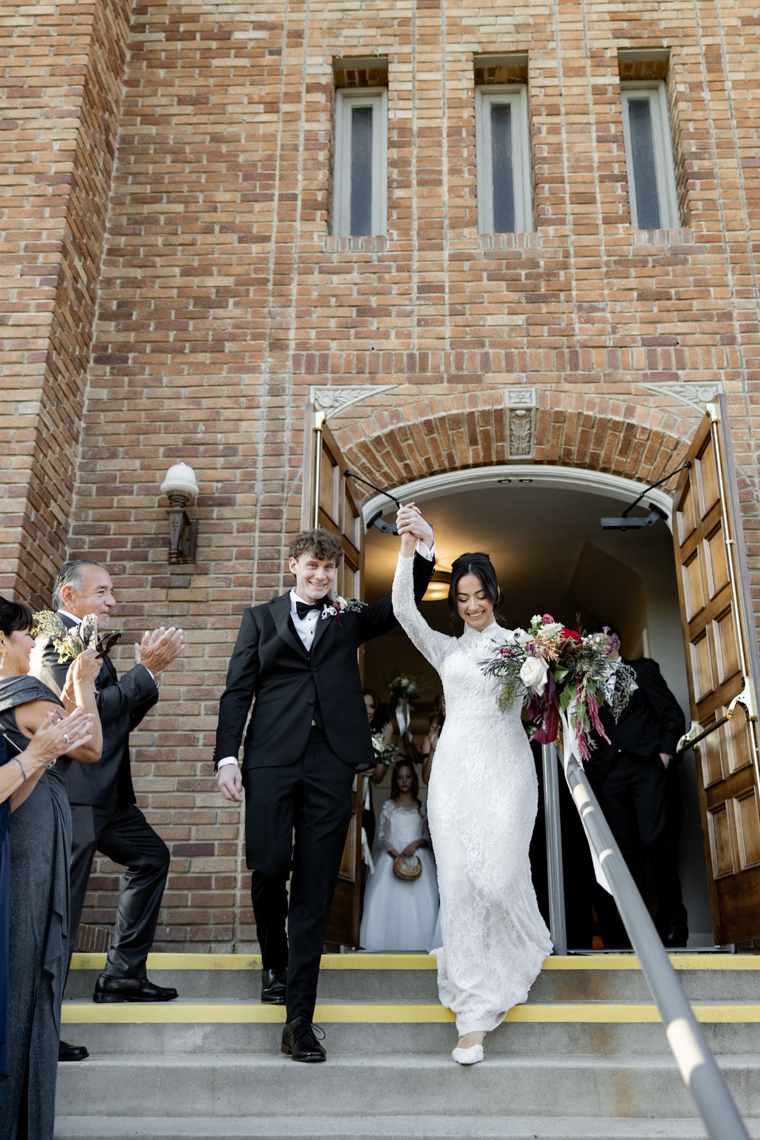 Newlyweds celebrate exit from brick church raising hands with bouquet as guests applaud.