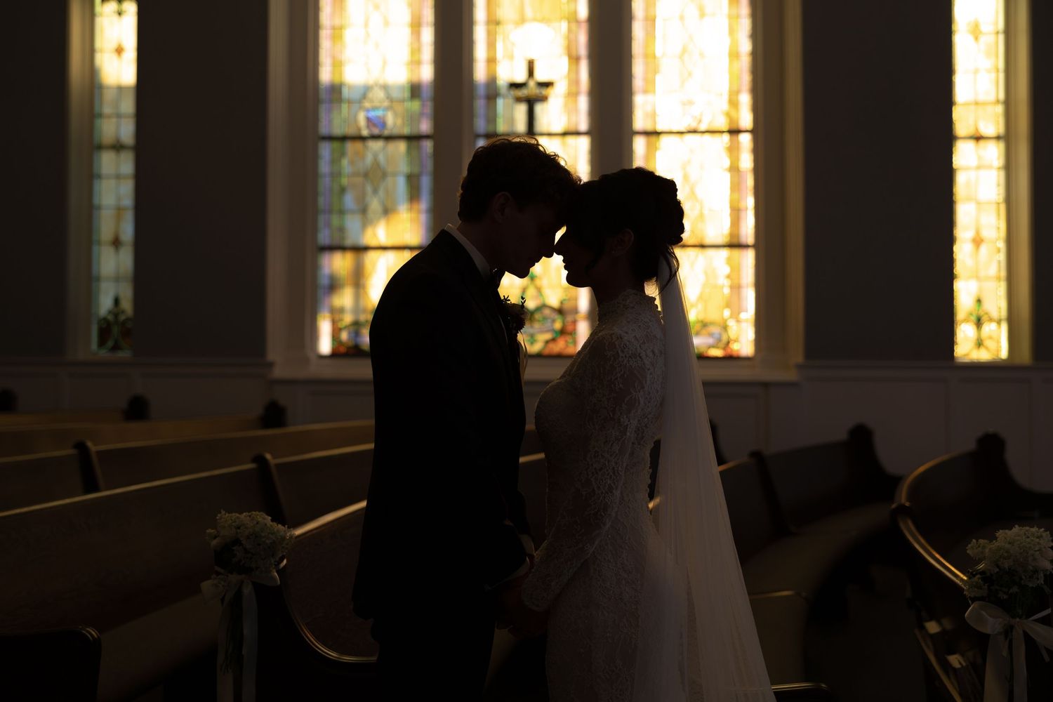 Silhouette of bride and groom touching foreheads in a church with glowing stained glass windows behind them.