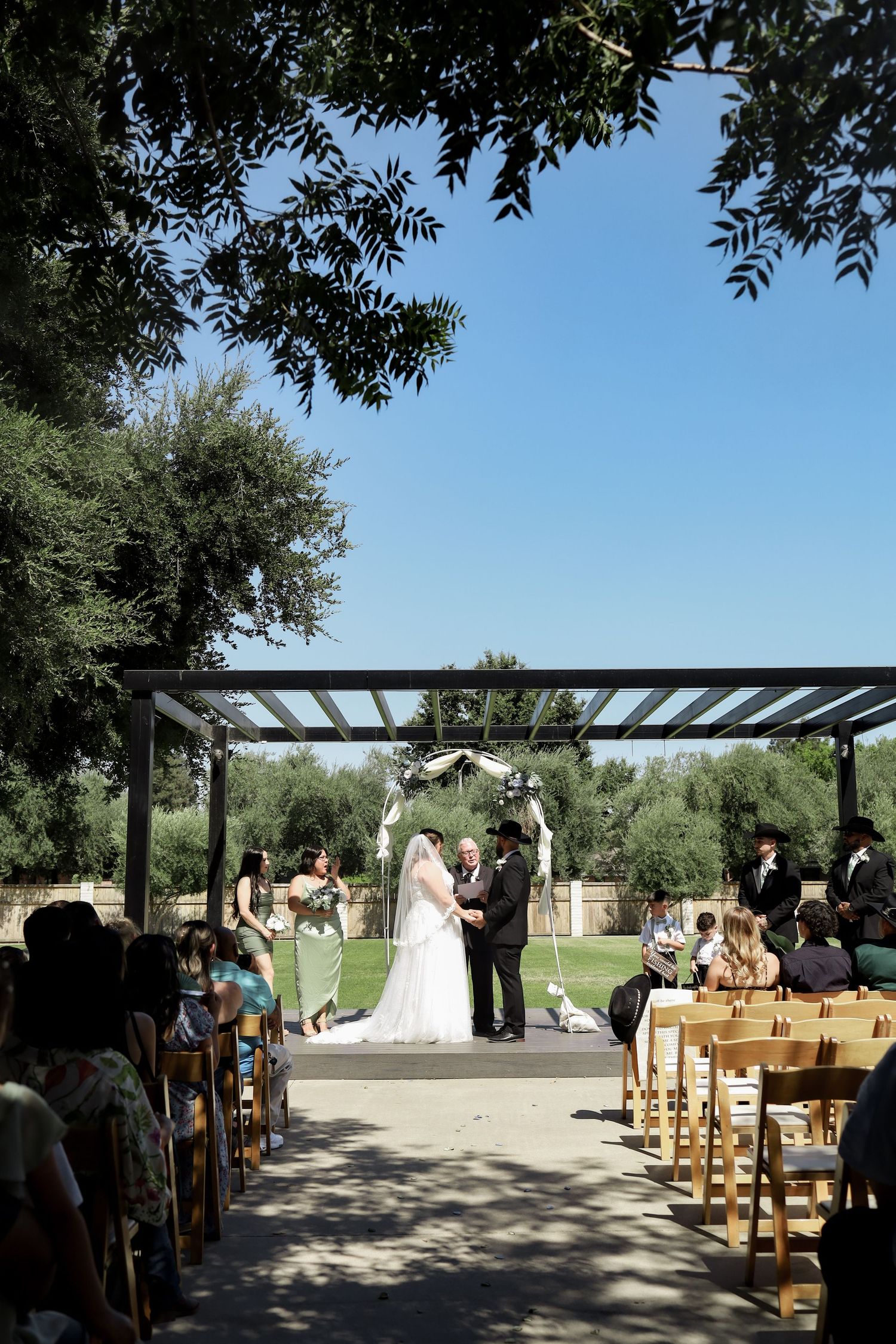An outdoor wedding ceremony under a pergola with wooden chairs and olive trees in a garden setting.