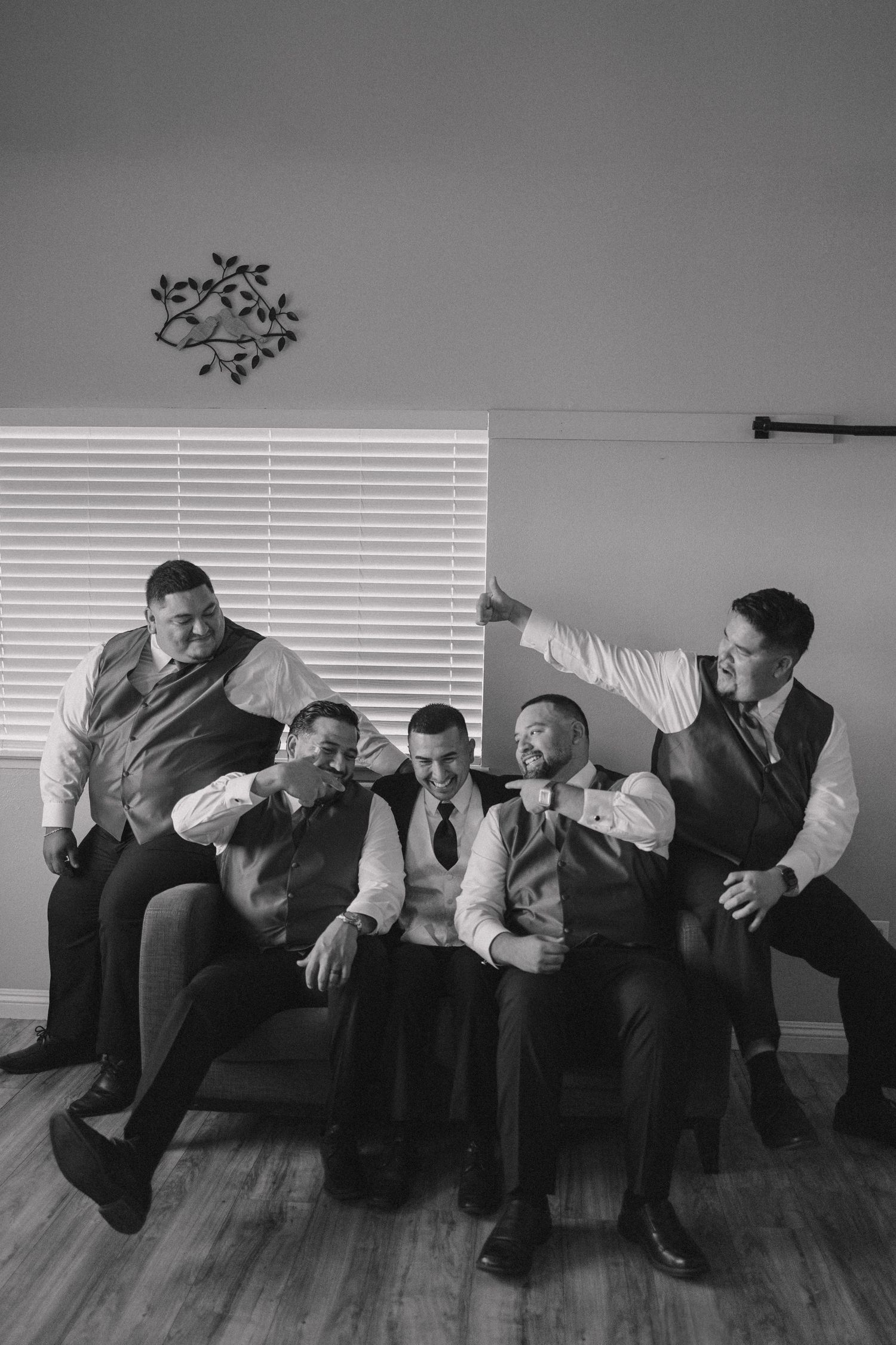 Playful groomsmen in formal attire posing together on a couch in black and white.