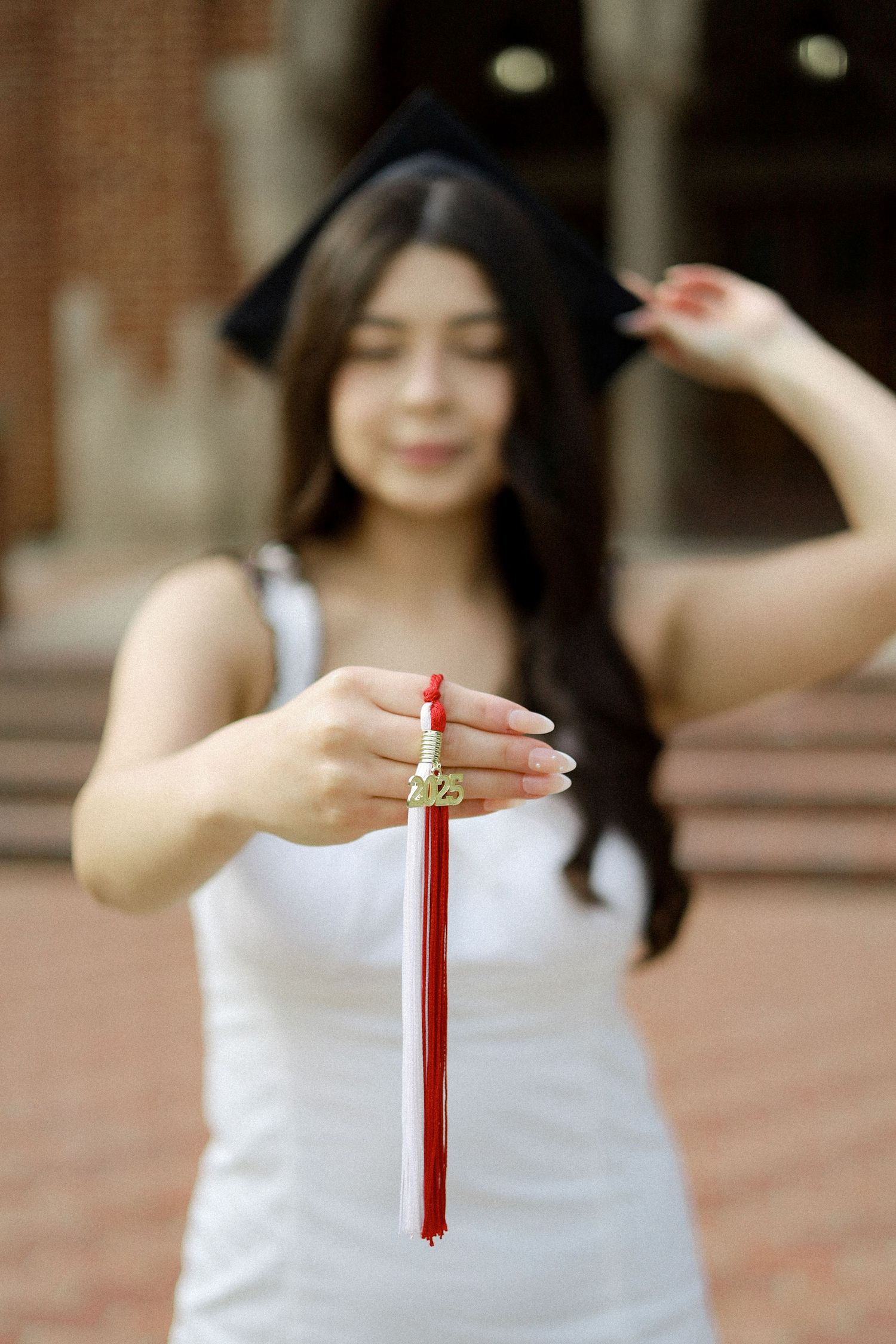 A graduate in white dress holds up a red graduation tassel while wearing a black cap during a graduation photoshoot.