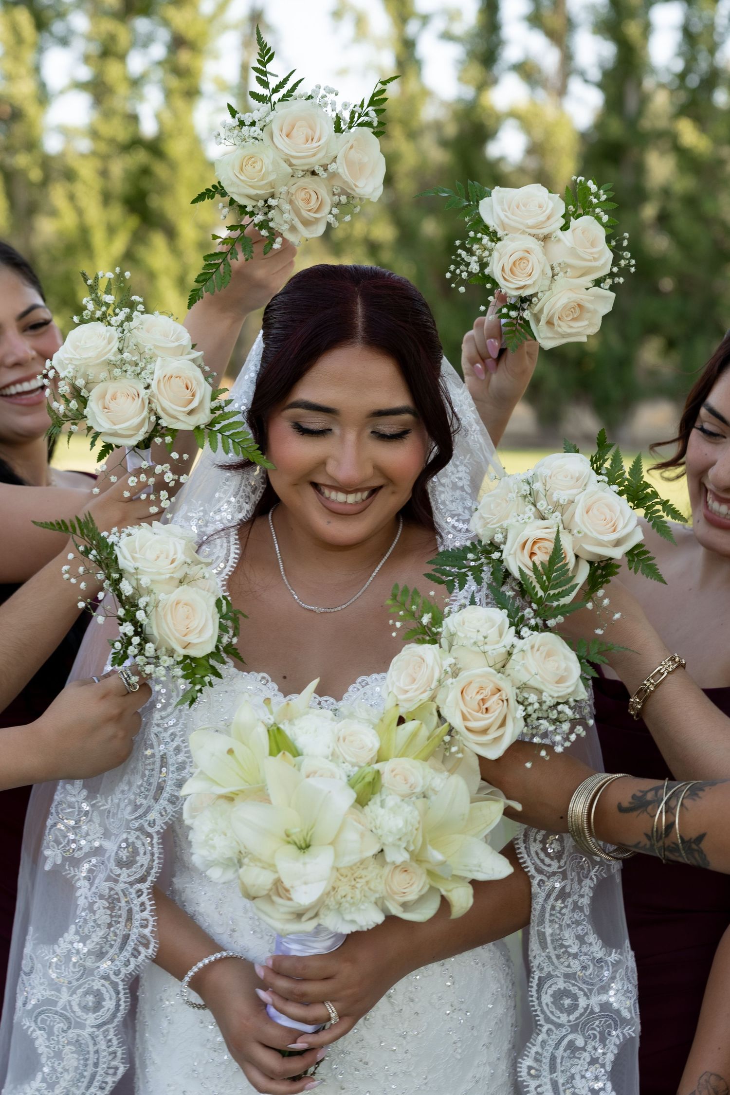 Smiling bride surrounded by bridesmaids holding white rose bouquets framing her face outdoors.