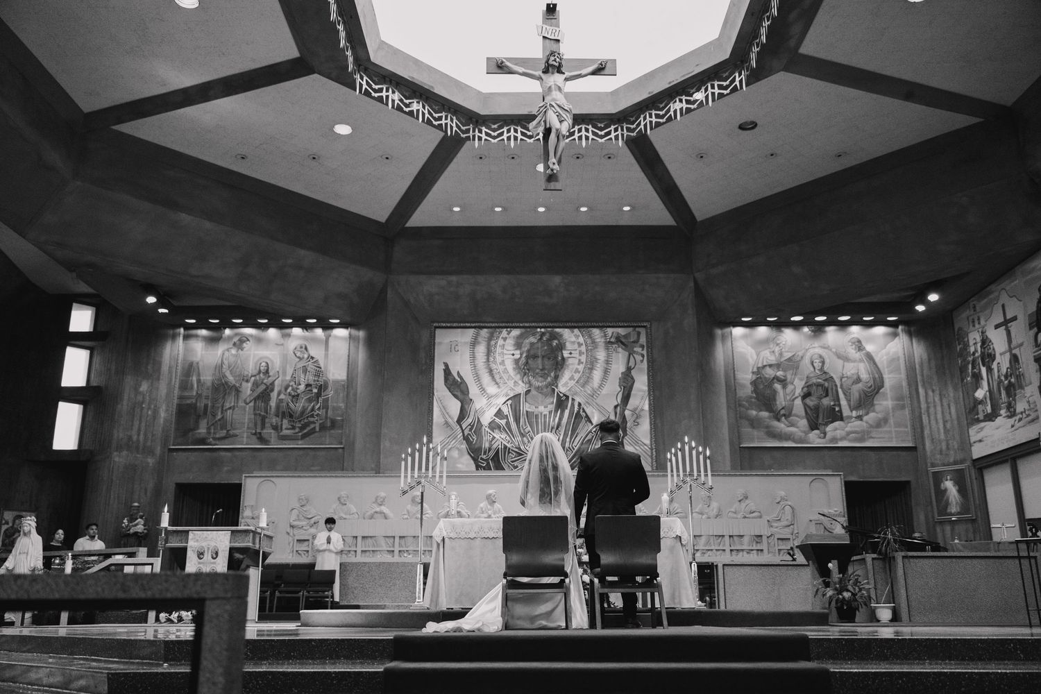 Black and white photo of a couple kneeling at a church altar during a wedding ceremony with religious murals.
