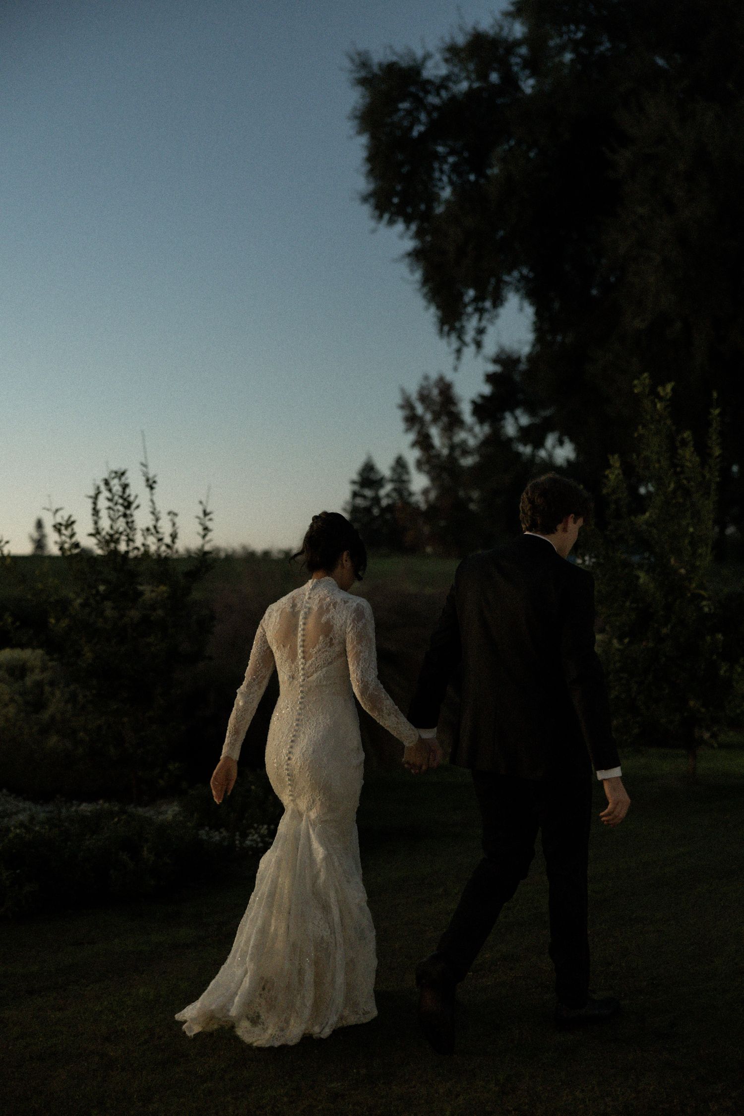 Bride and groom holding hands walking through garden at dusk in elegant lace wedding gown and suit.
