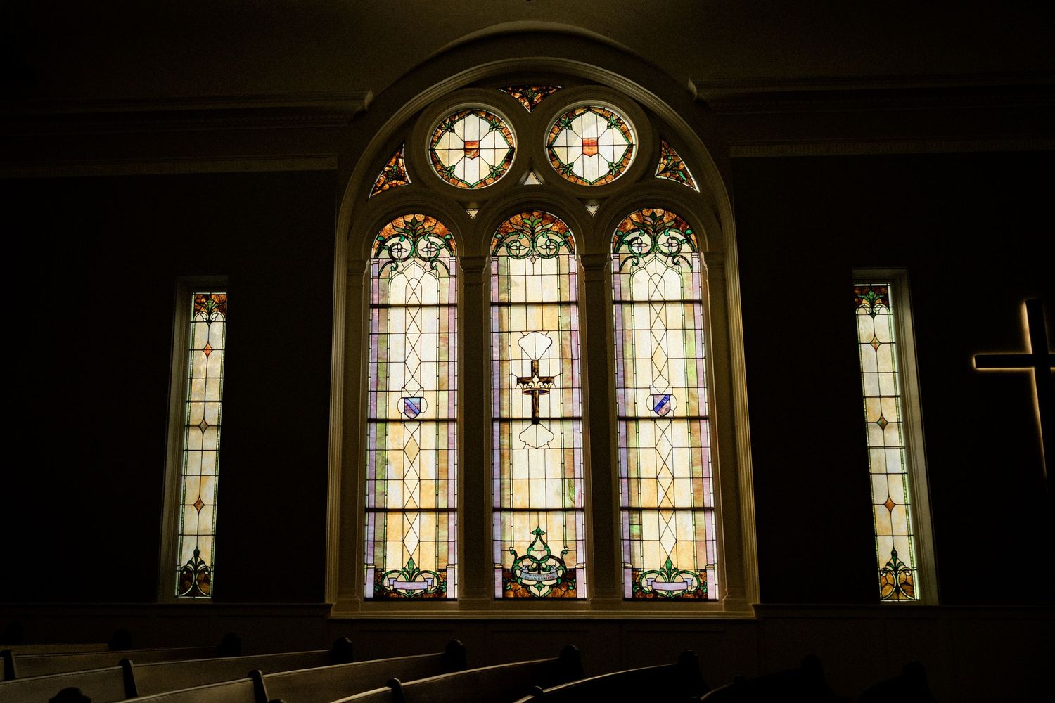 Ornate stained glass church windows featuring a cross design, flanked by colorful panels, illuminated in a dark sanctuary with wooden pews.