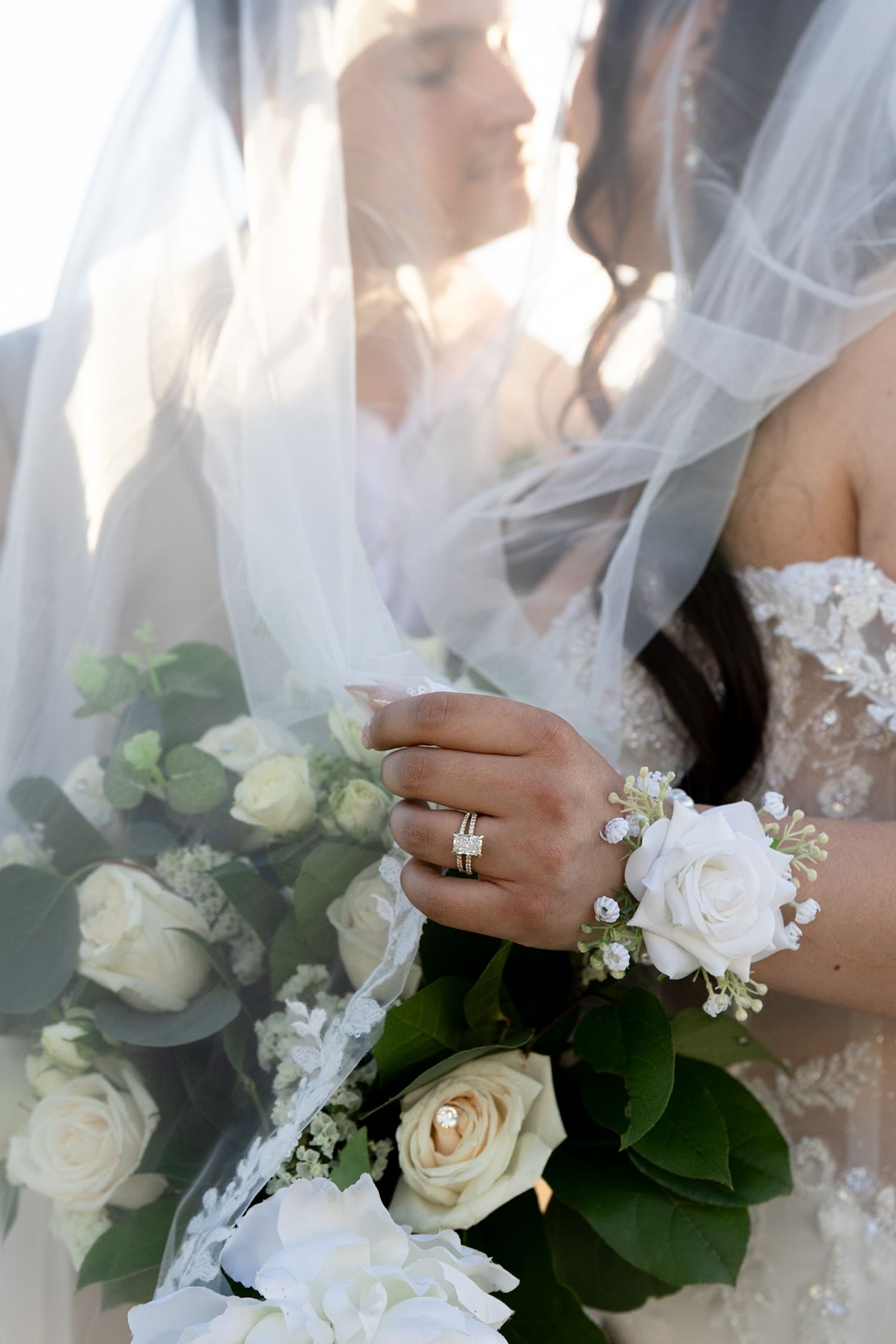Bride holding white rose bouquet with wedding ring and floral wrist corsage, veil draped over couple kissing.