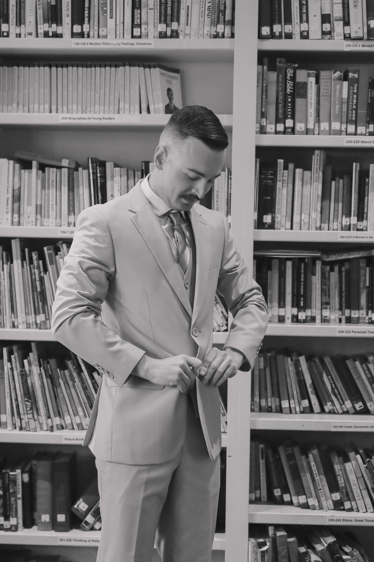 A groom in a light gray suit checks his watch while standing in a library.