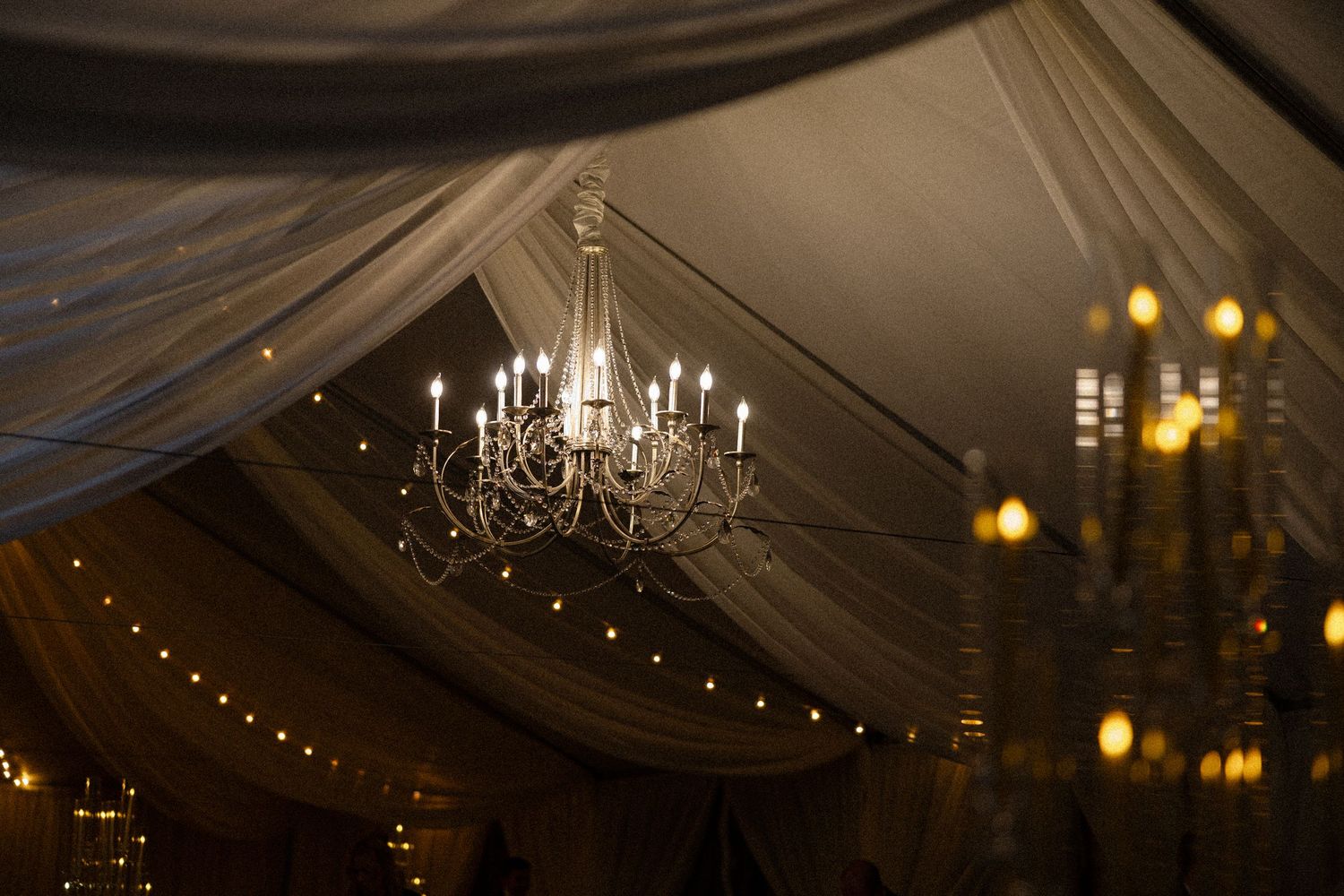 Elegant crystal chandeliers and string lights illuminate a draped wedding reception tent ceiling.
