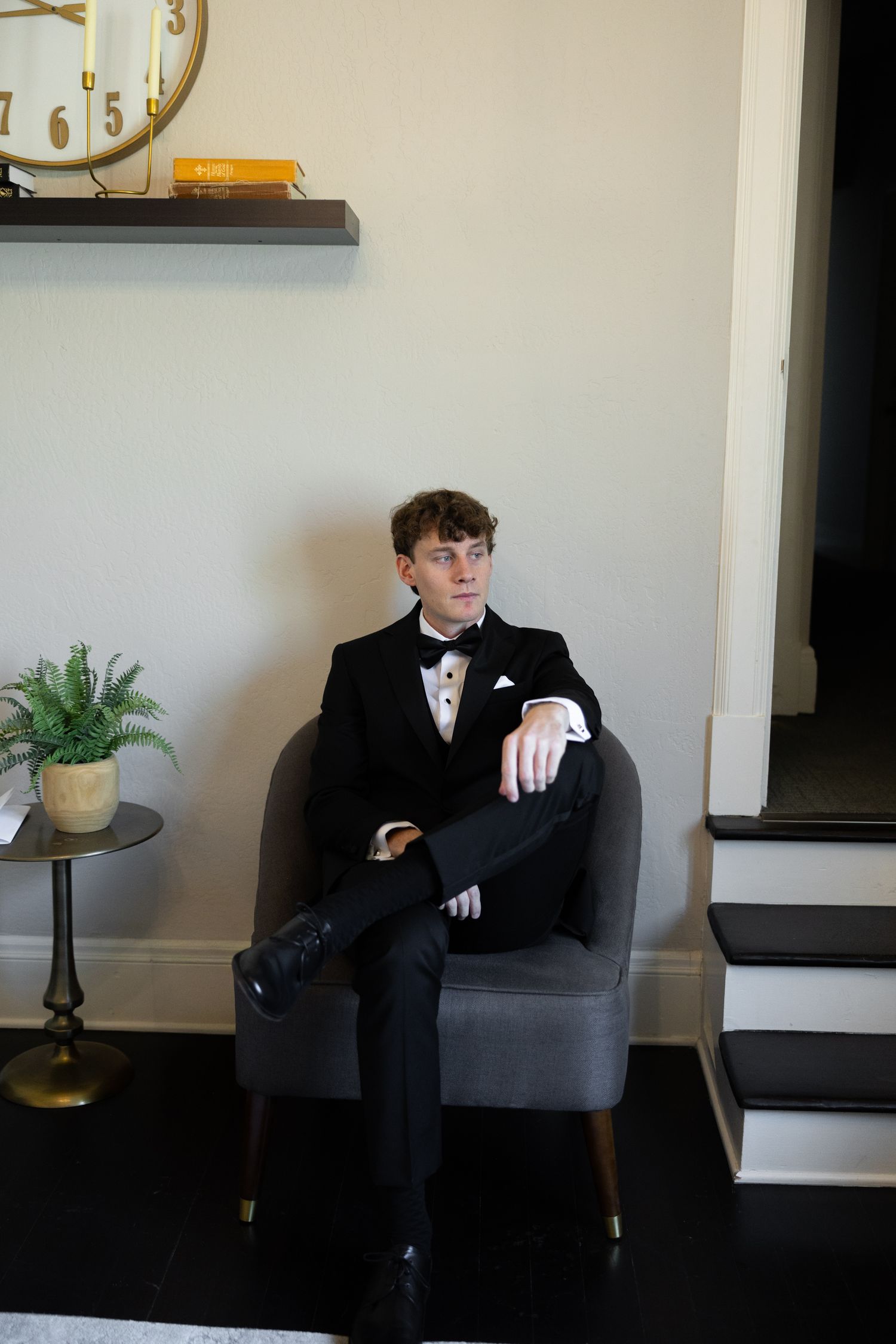 Young man in black tuxedo with bow tie seated in gray chair in elegant room.