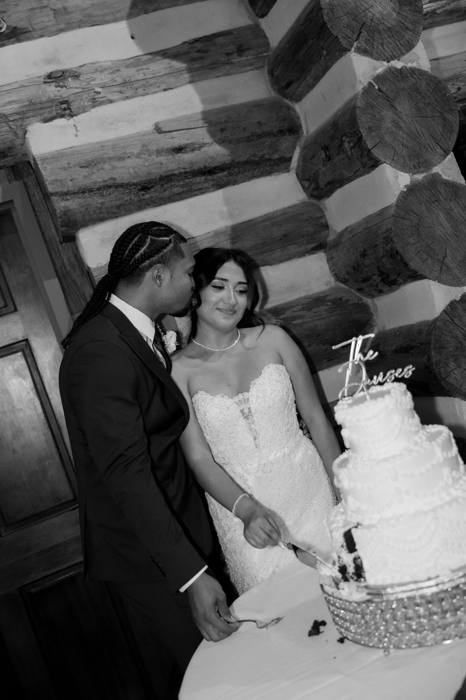 Black and white photo of bride and groom cutting tiered wedding cake in rustic log cabin.