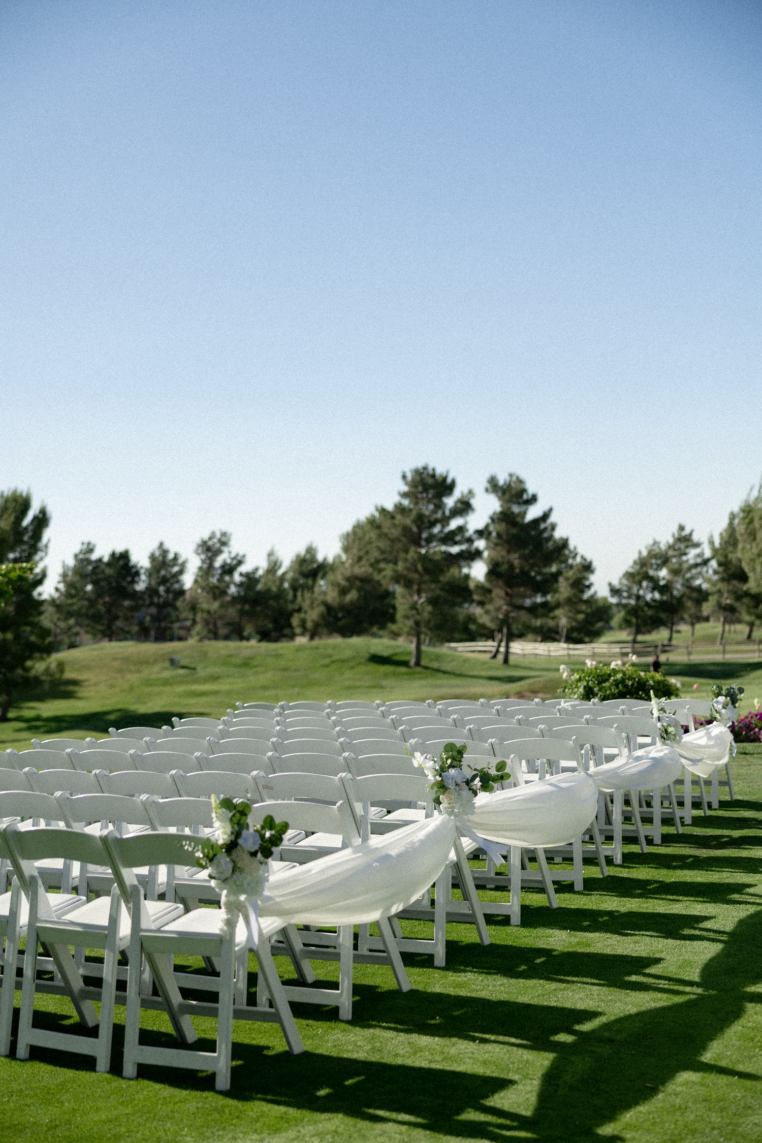 White folding chairs arranged in rows on lush green grass for an outdoor wedding ceremony under a clear blue sky.