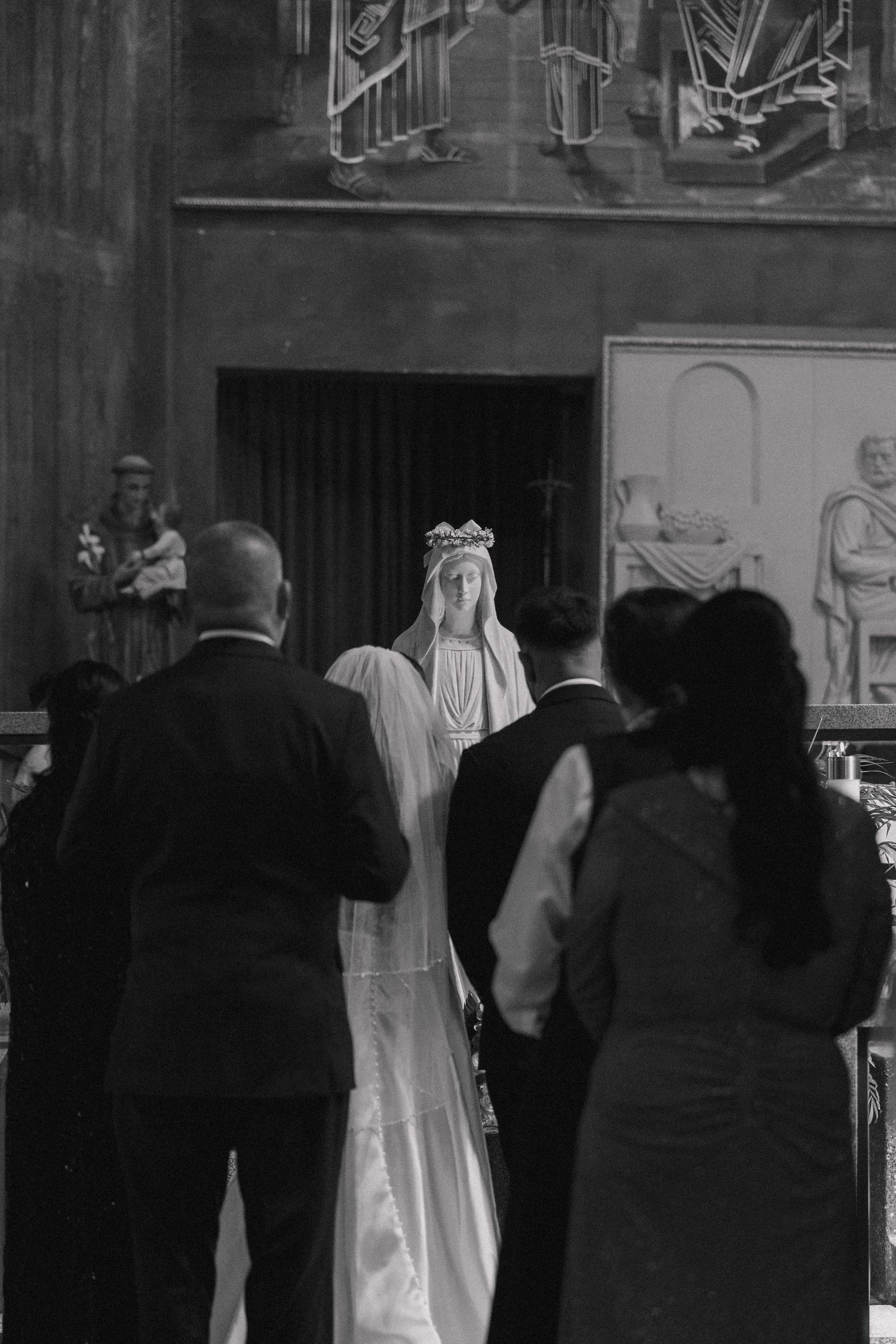 Black and white photo of people praying before a Virgin Mary statue inside a Catholic church.