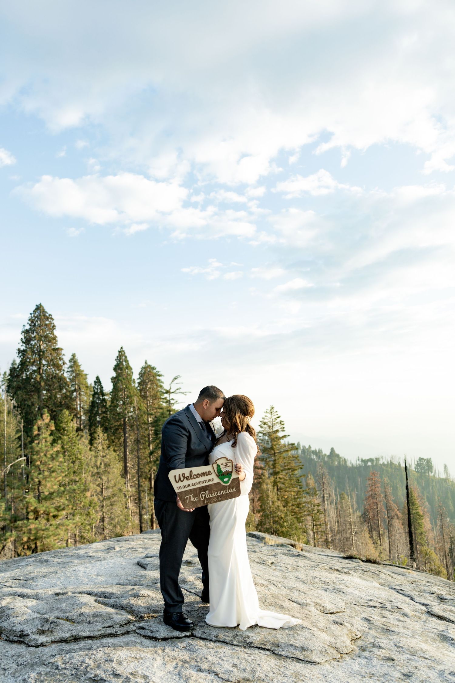 Couple shares intimate moment on scenic mountain overlook with evergreen trees and cloudy sky in background.