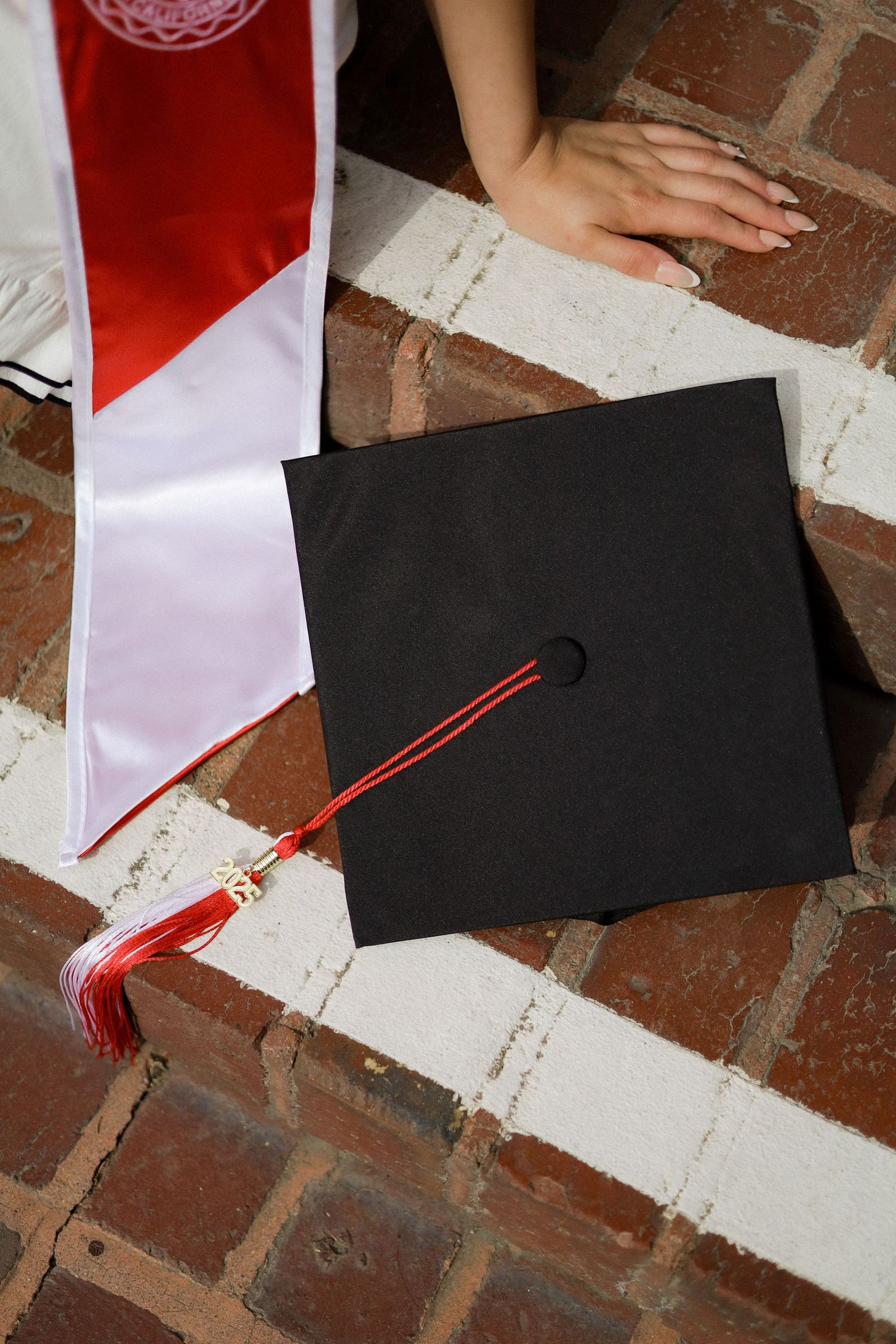 A graduation cap with red tassel being placed on a brick surface next to white paper.