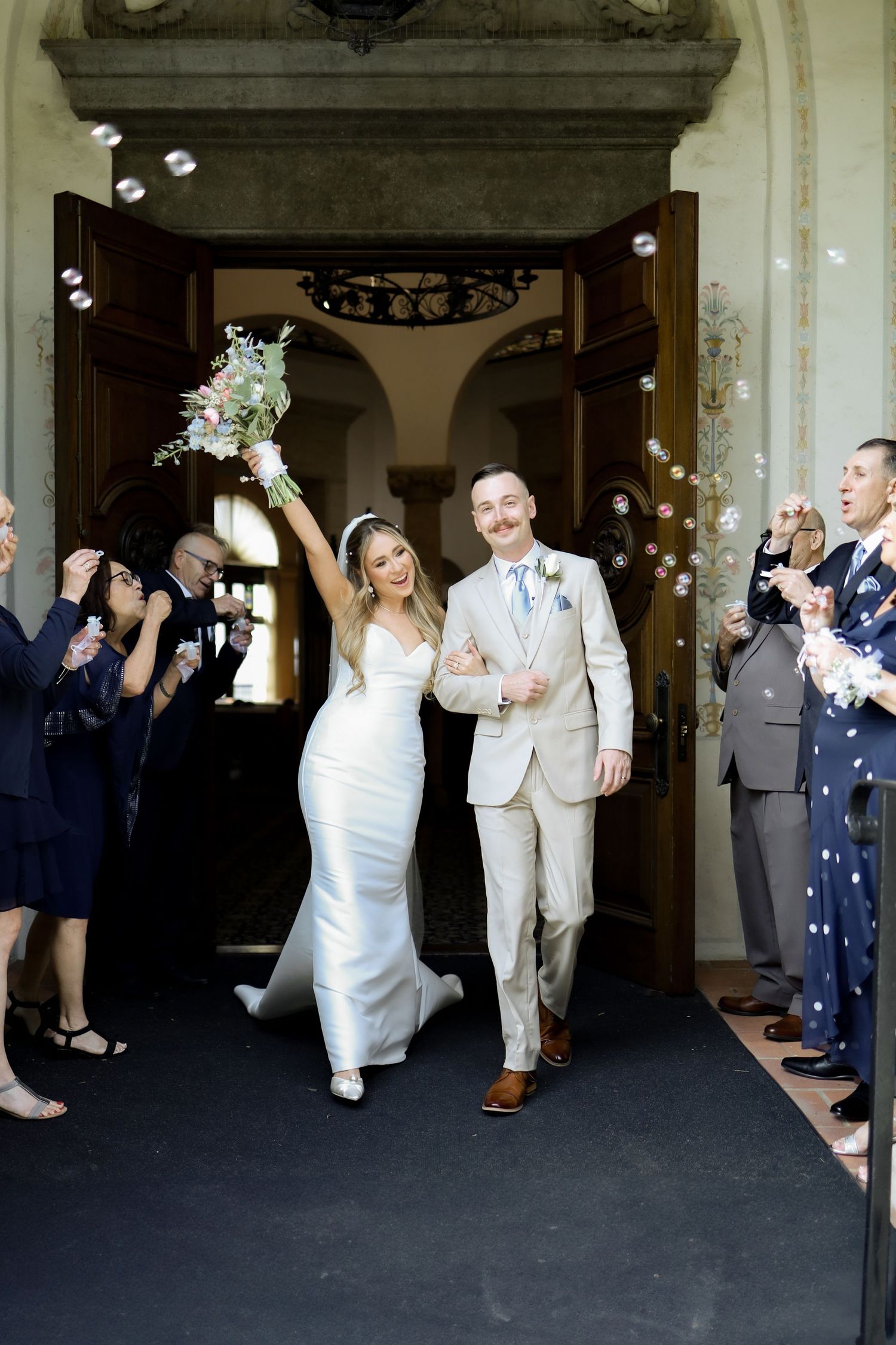 Newlyweds exit church while guests throw flower petals in celebration.