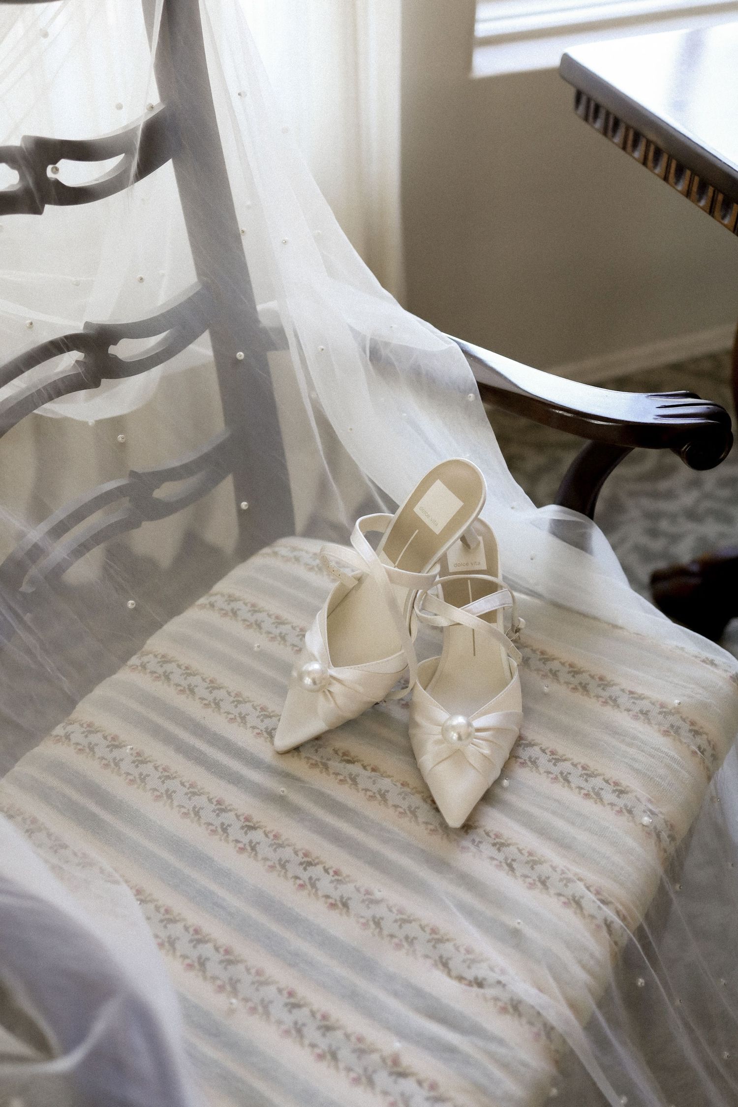 Elegant cream-colored bridal shoes displayed on a patterned cushion with veil overlay.