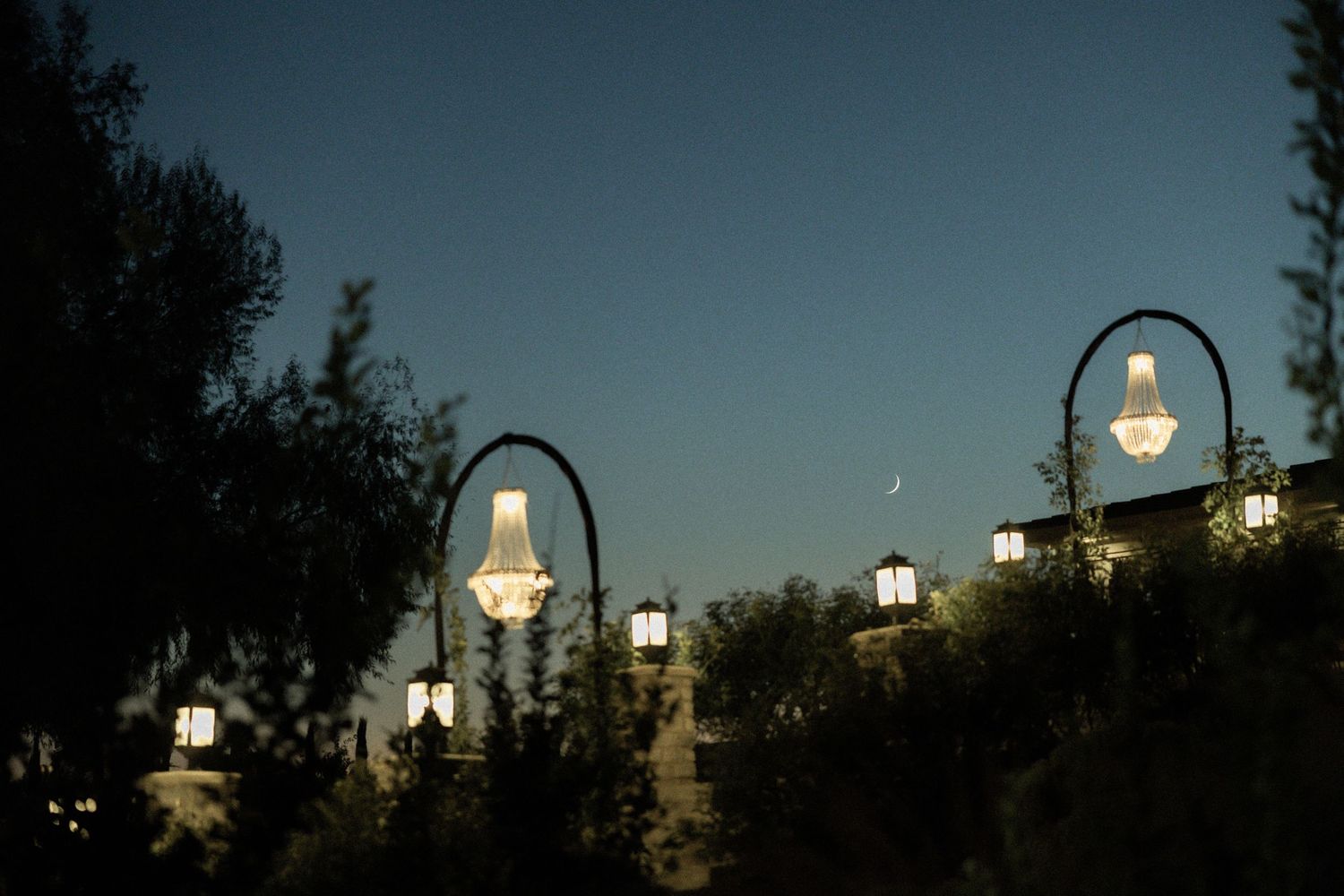 Elegant outdoor chandelier lanterns glow against twilight sky with crescent moon visible.