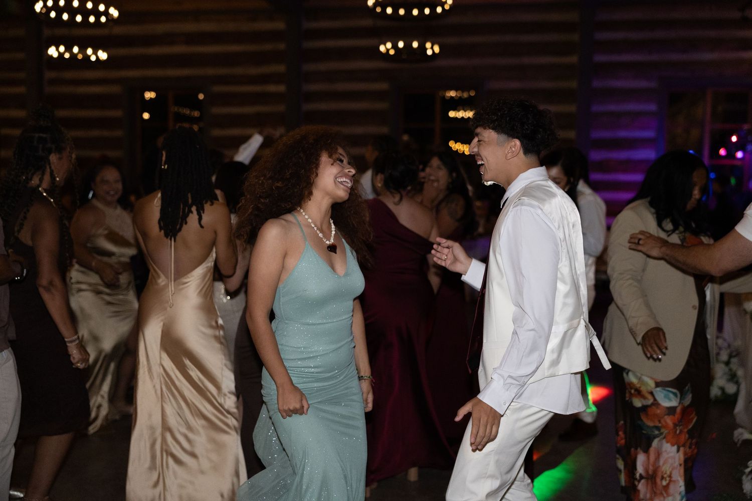 Teens dancing together at a prom or formal event in elegant attire under warm string lights.