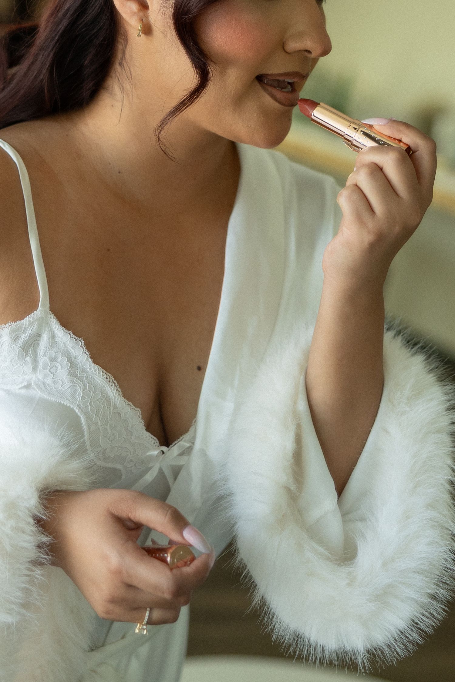 A bride in white lace lingerie and feather robe applies pink lipstick while holding the gold tube case.