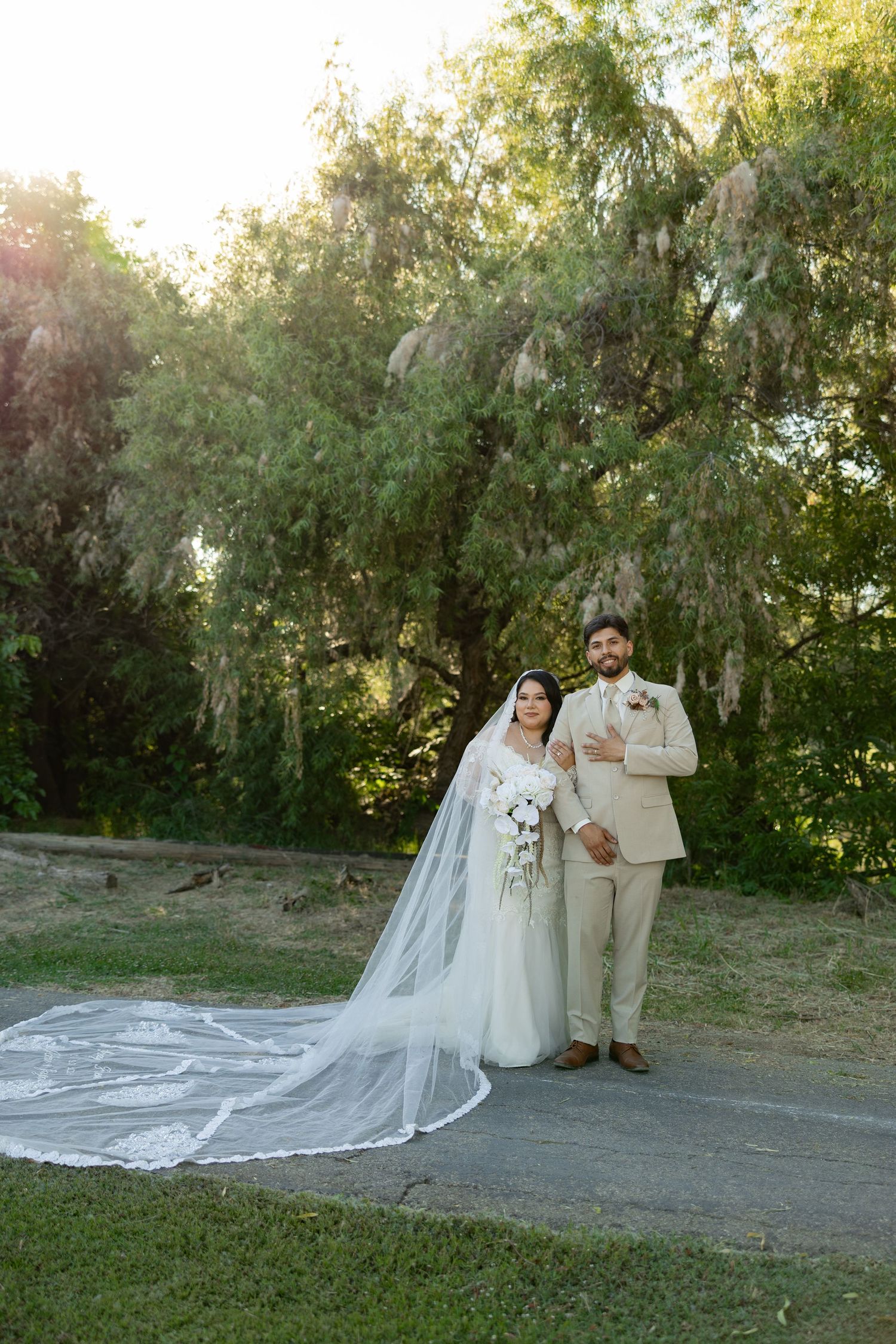 Bride in white gown with long veil and groom in tan suit pose together outdoors under lush green trees on their wedding day.