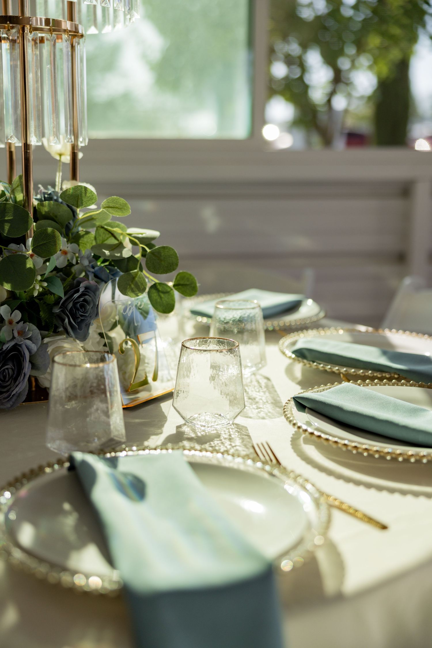Elegant wedding table setting with gold chargers, teal napkins, crystal glasses, and floral centerpiece in soft natural light.