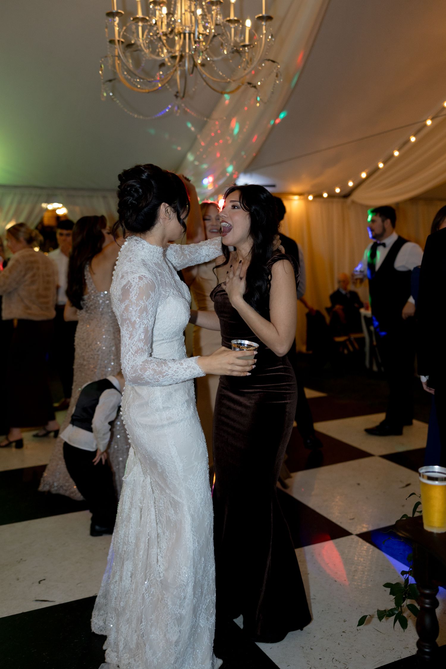 Bride in lace gown dances and laughs with female guest in black dress at wedding reception.