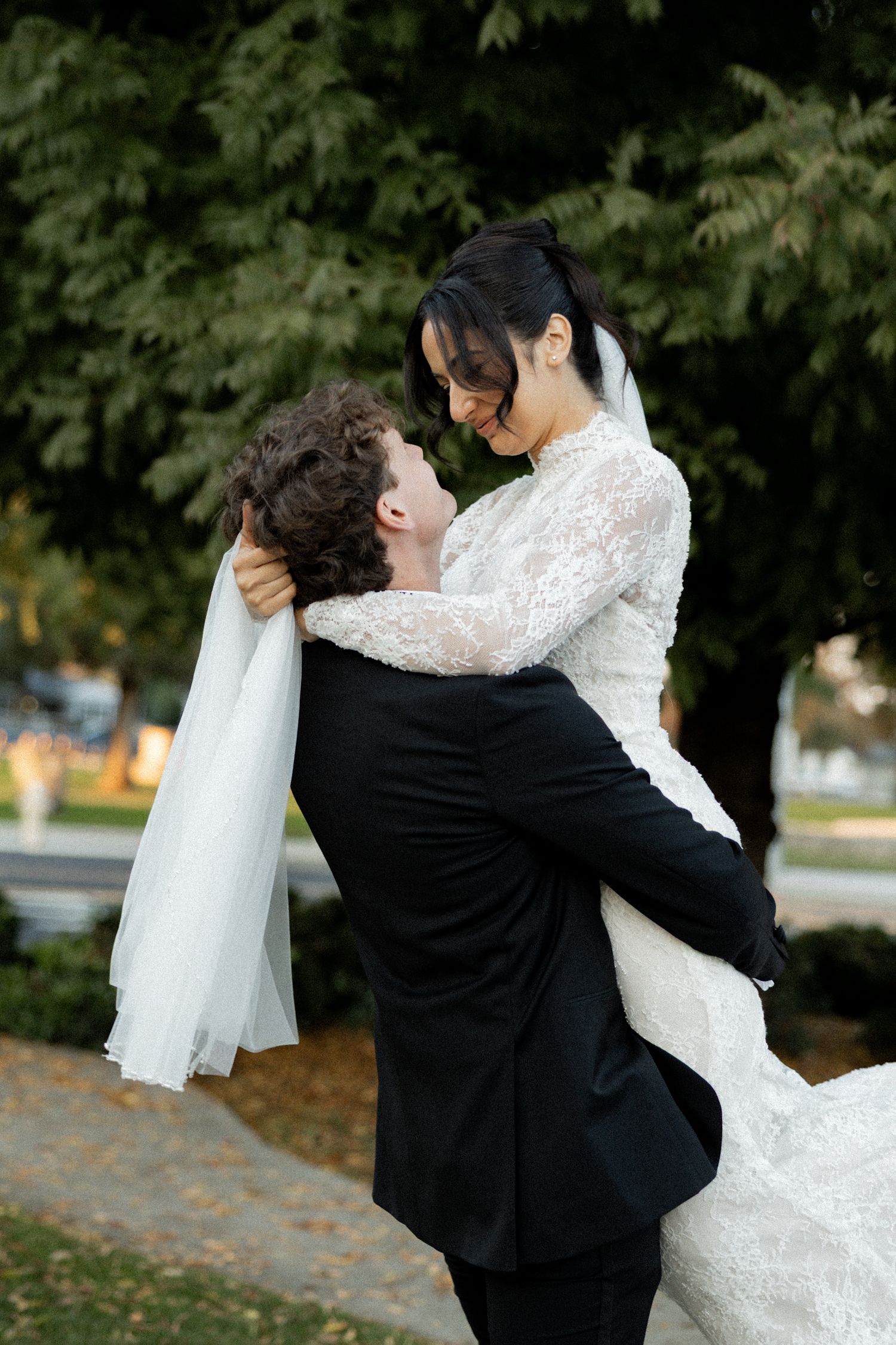 Groom lifting smiling bride in lace wedding dress outdoors surrounded by green trees.