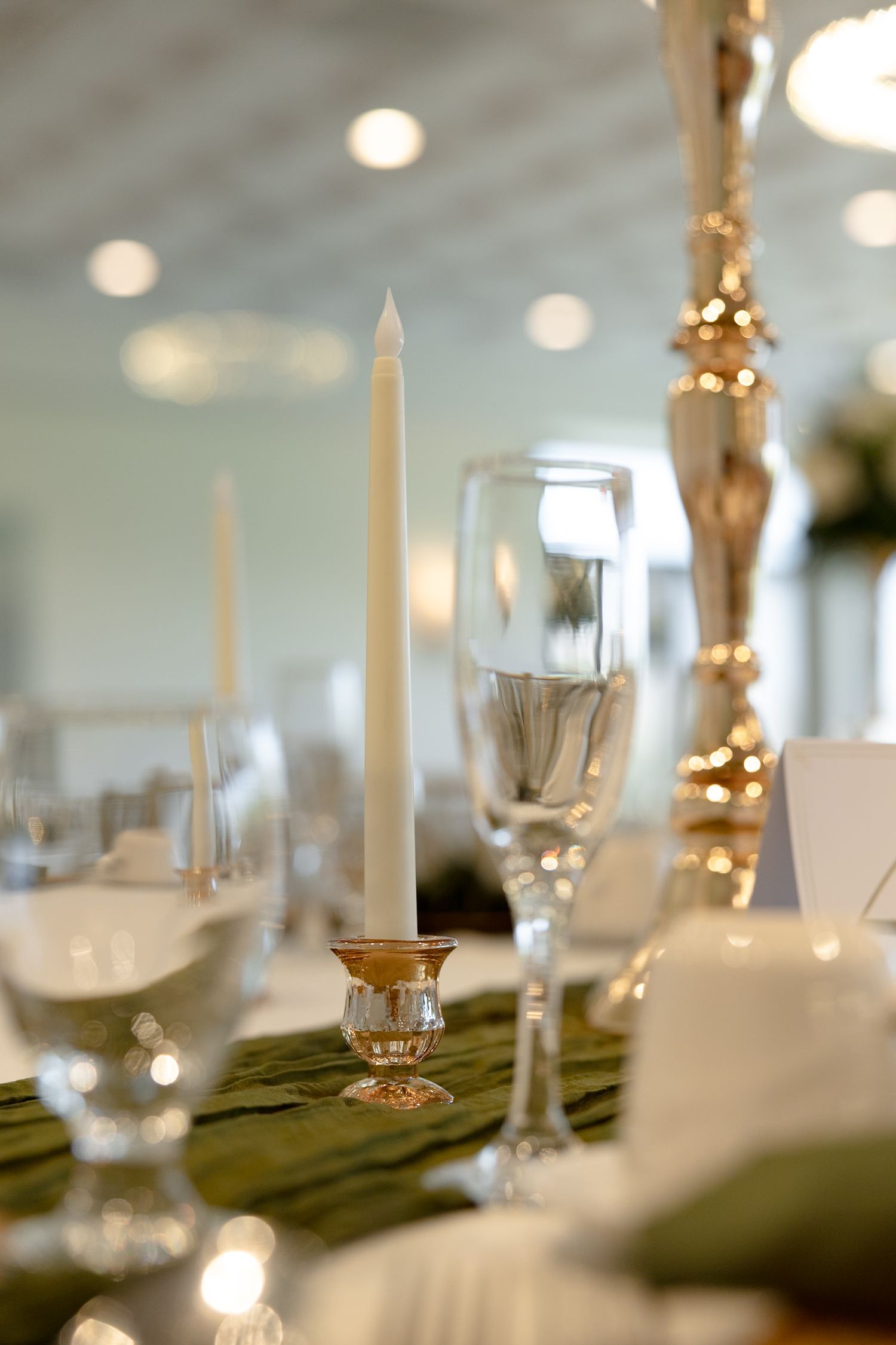 Elegant wedding table setting with white taper candles, crystal champagne flutes, and green table runner in a bright reception hall.
