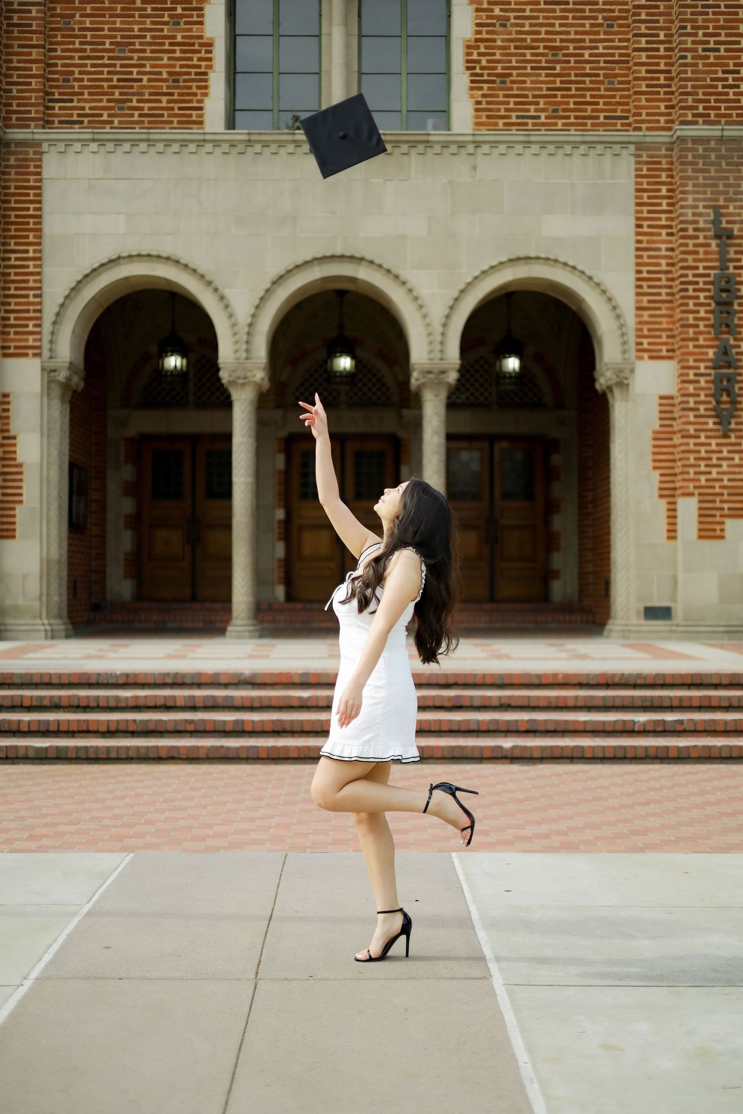 Graduate in white dress and cap poses playfully in front of brick university building with arches.