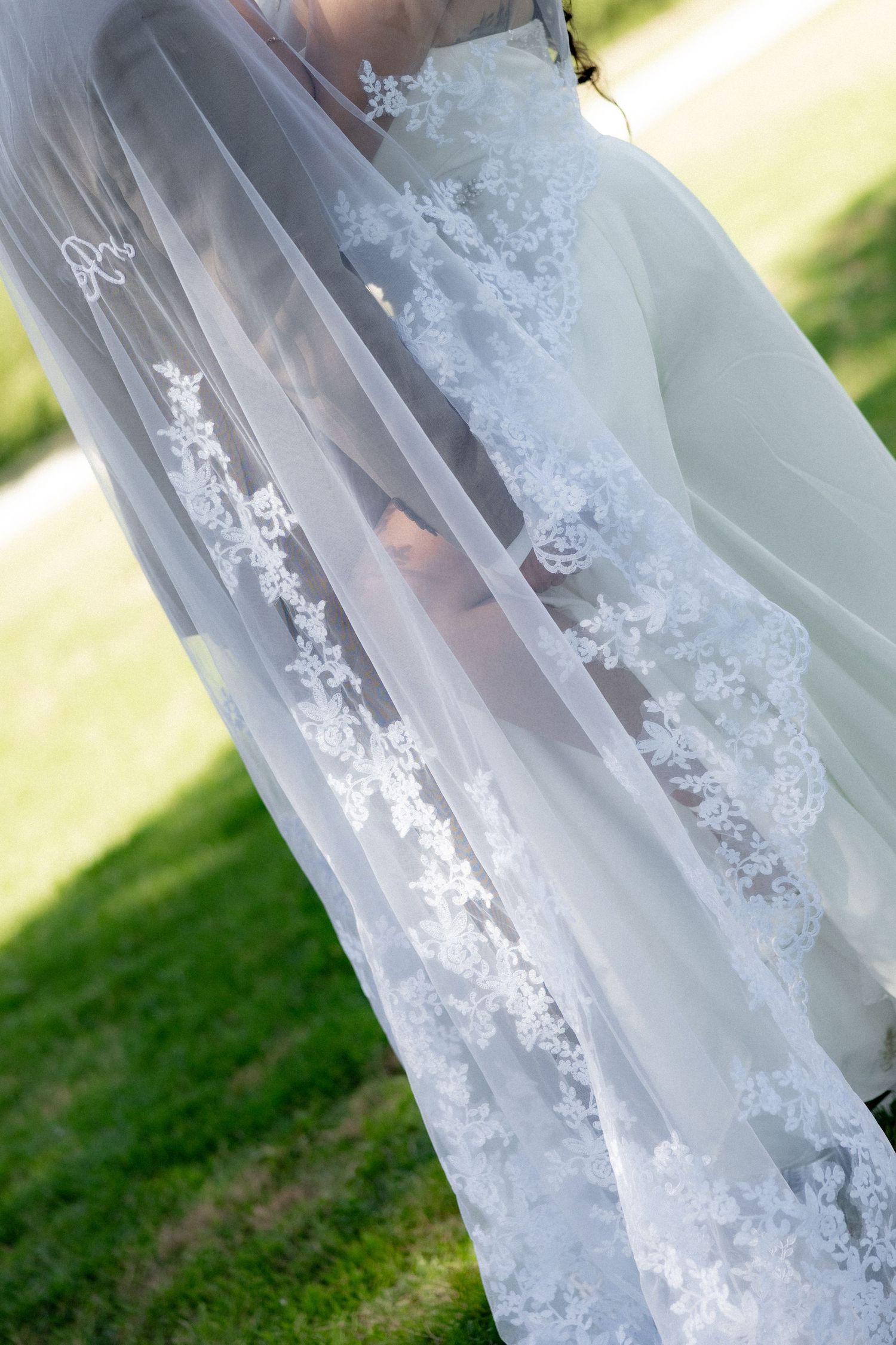Bride in white wedding dress with elegant lace-trimmed veil outdoors on green grass.
