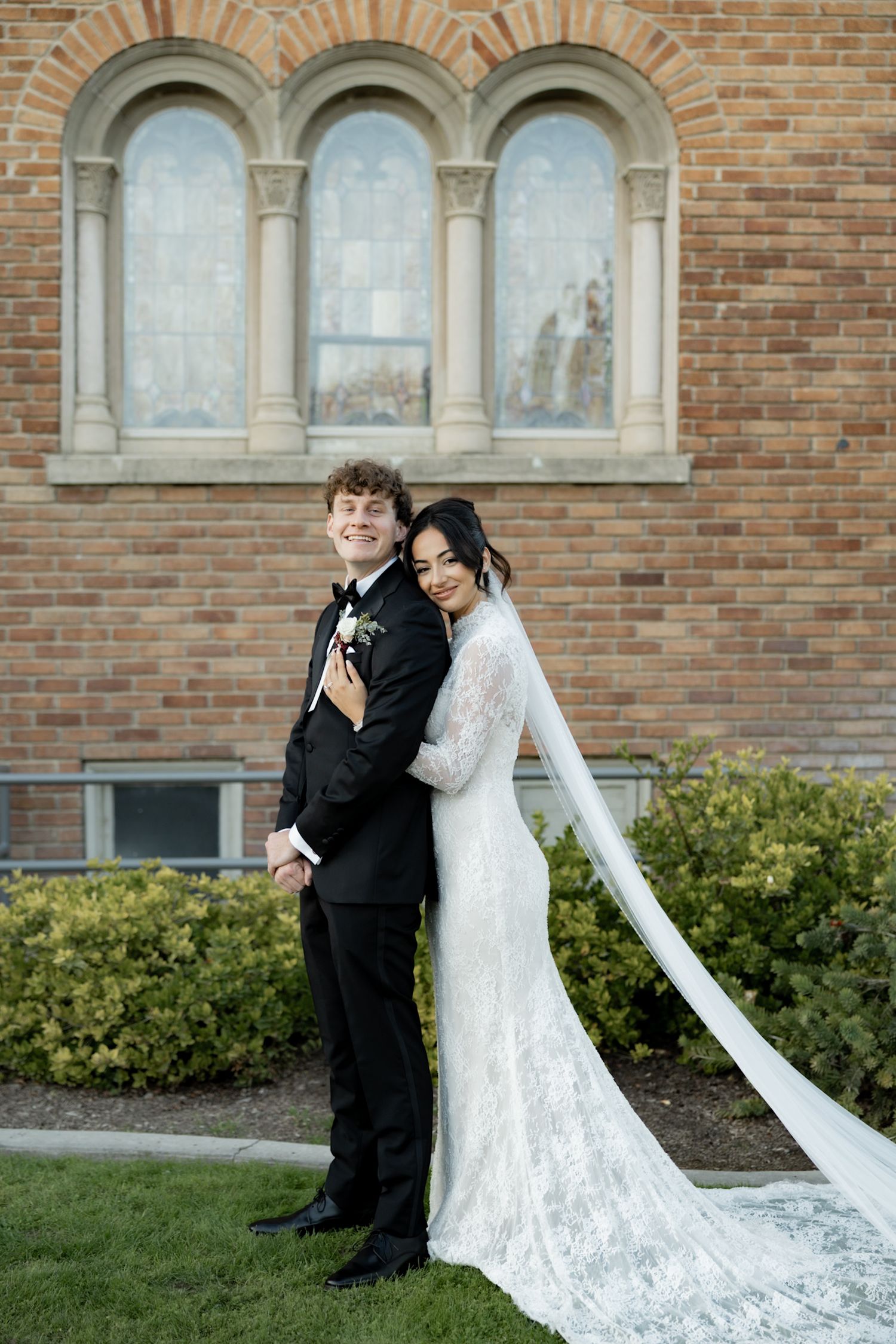 Bride and groom pose outside brick church, bride in lace gown with cathedral veil.