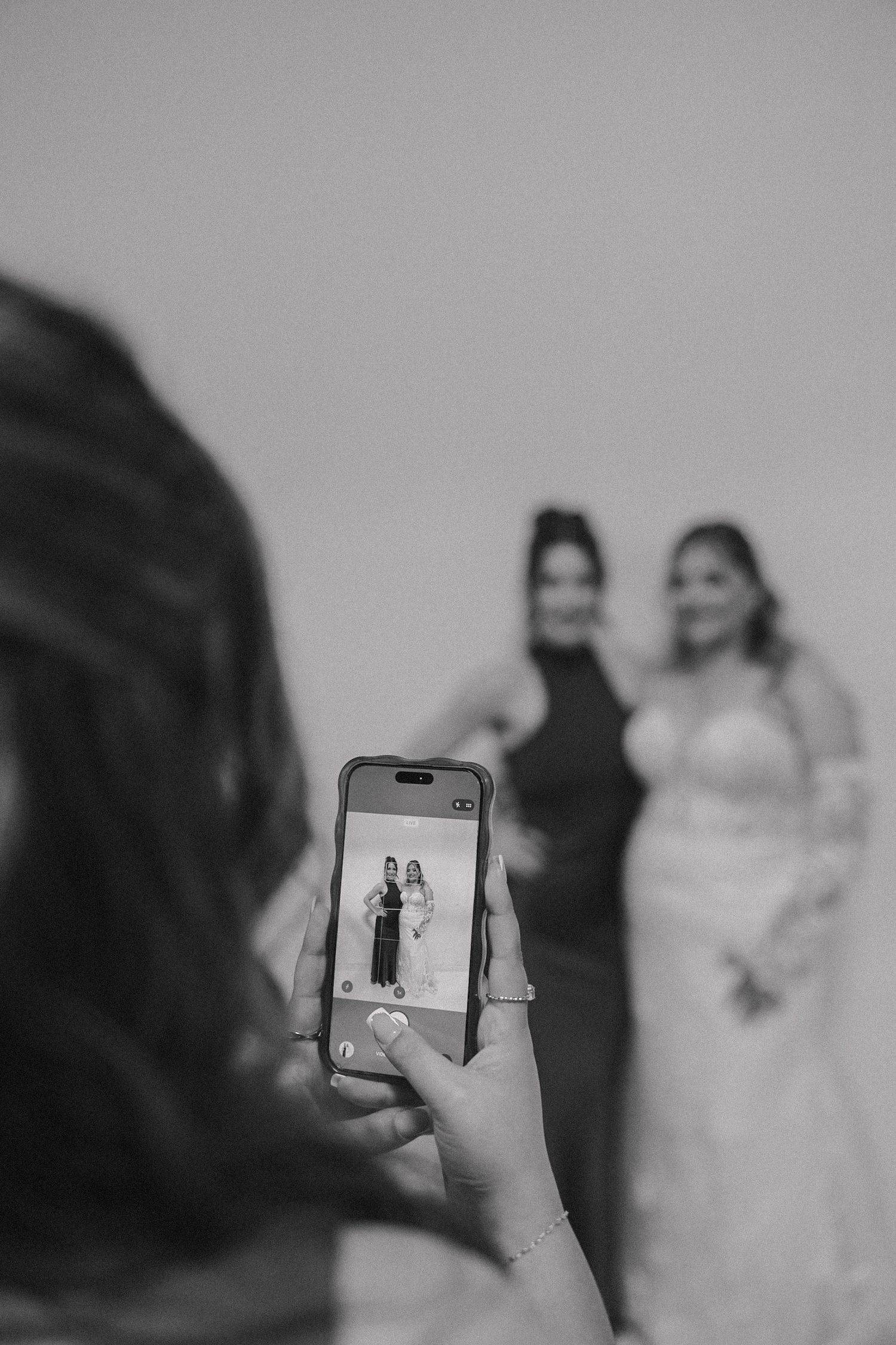 Black and white photo of a person photographing two women posing together, one in a wedding dress, captured on a smartphone screen.