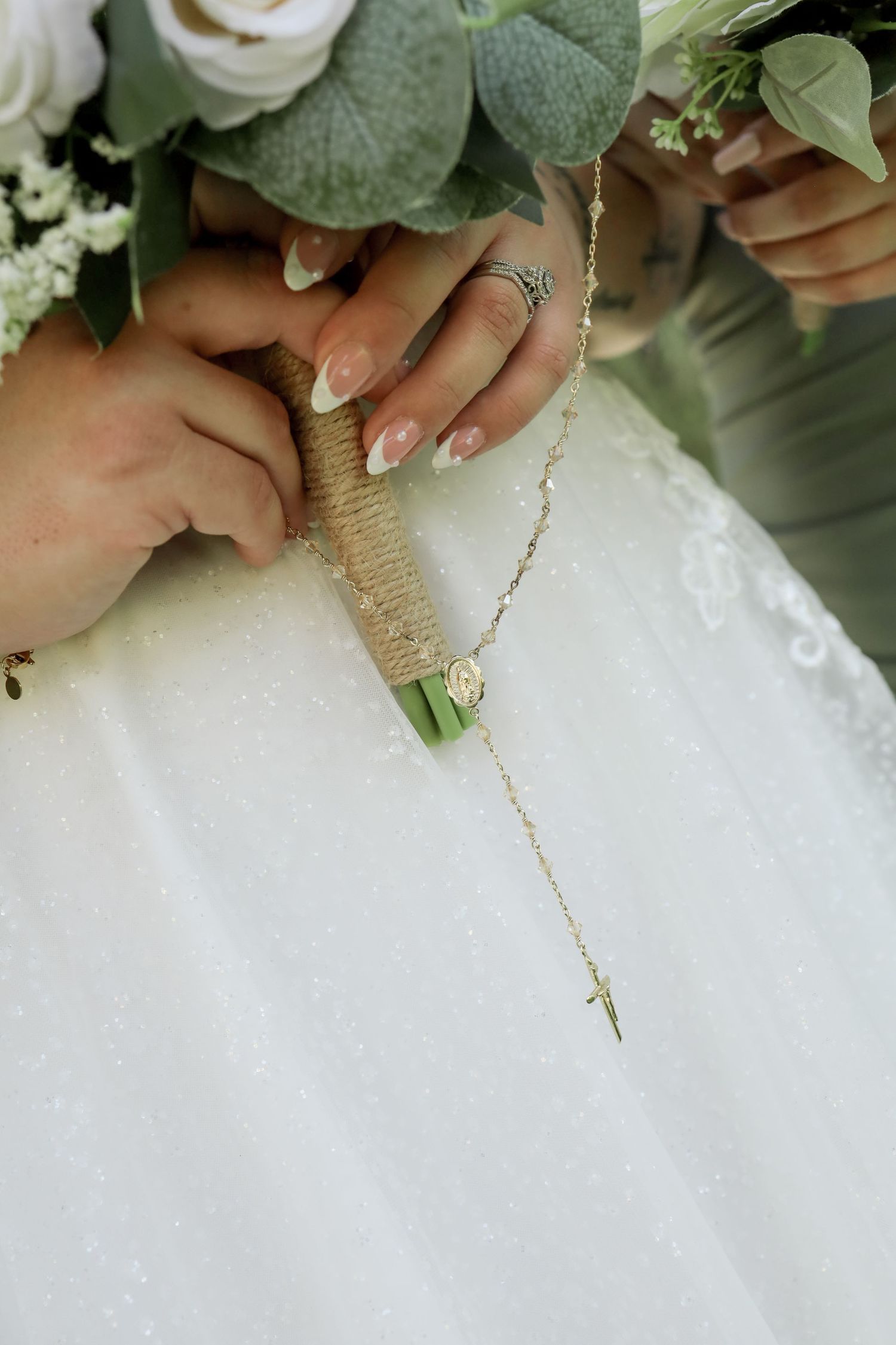 Close-up of bridal bouquet wrapped in twine with a delicate necklace and manicured nails against white dress fabric.