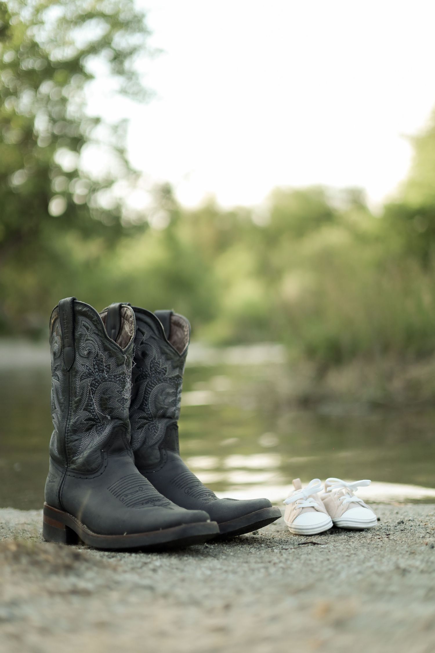 A pair of black cowboy boots and white sneakers arranged on a concrete surface near water.