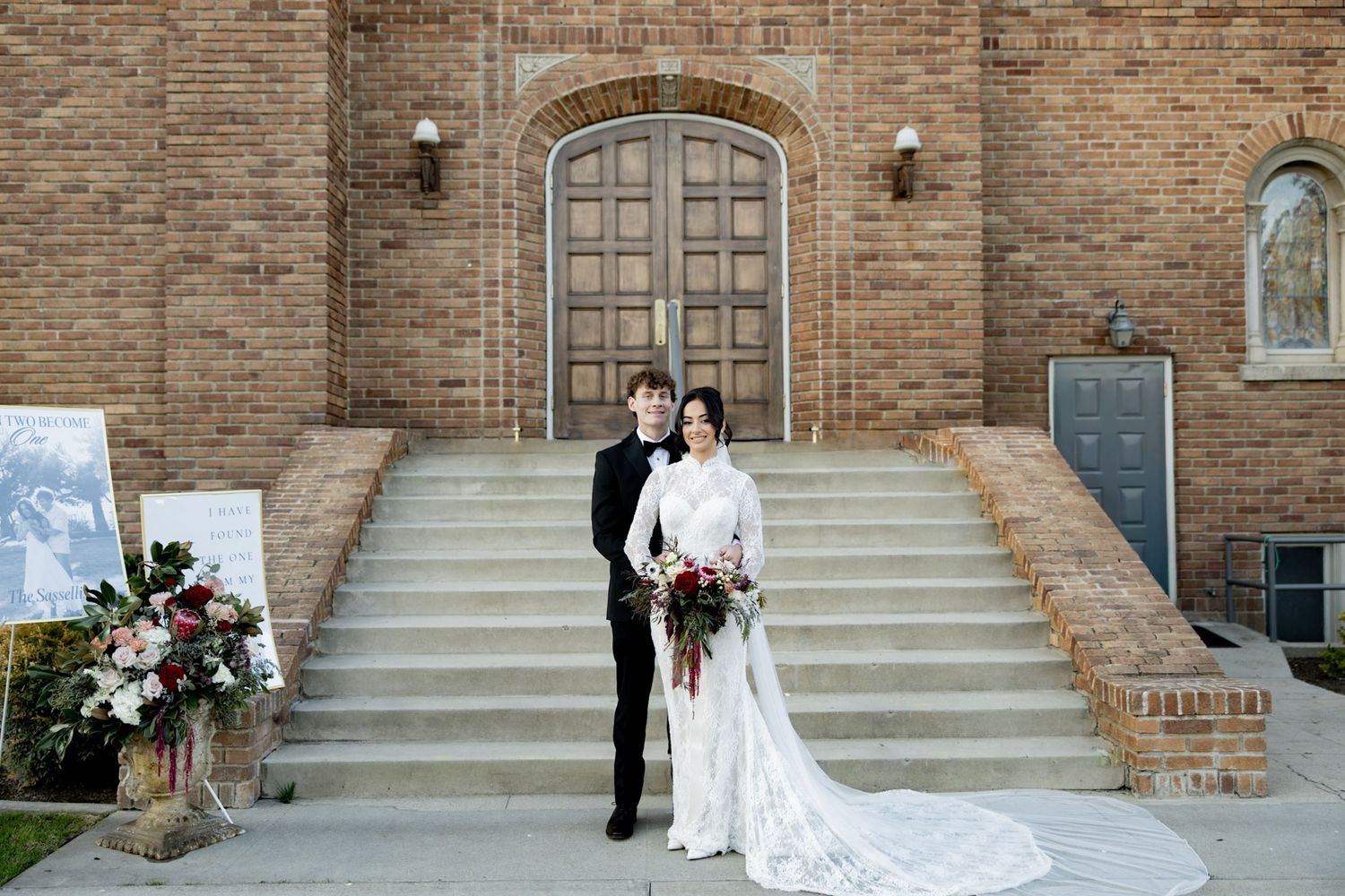 Bride and groom posing on church steps in elegant wedding attire with floral arrangements nearby.