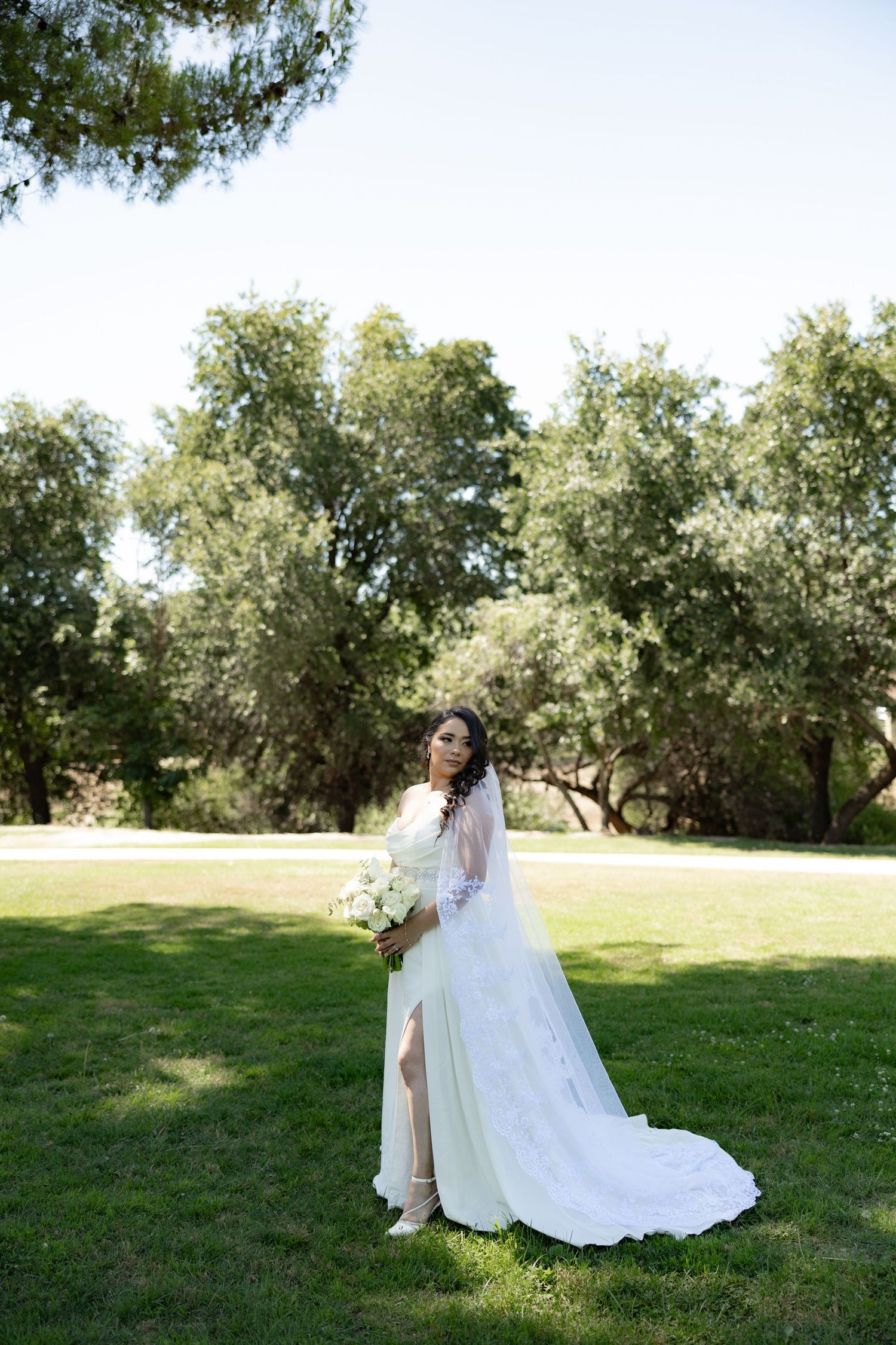 Bride in white lace gown with veil holds bouquet on green lawn surrounded by trees on sunny day.