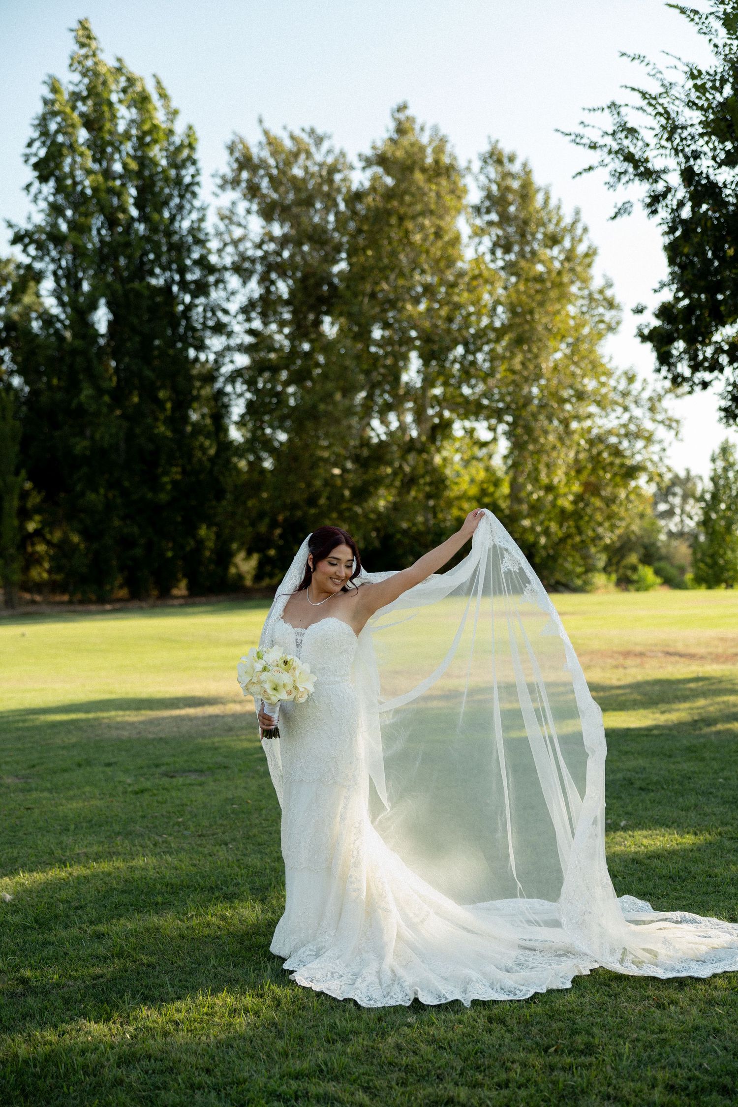 Bride in lace wedding gown holding veil and bouquet on green lawn with trees in background.