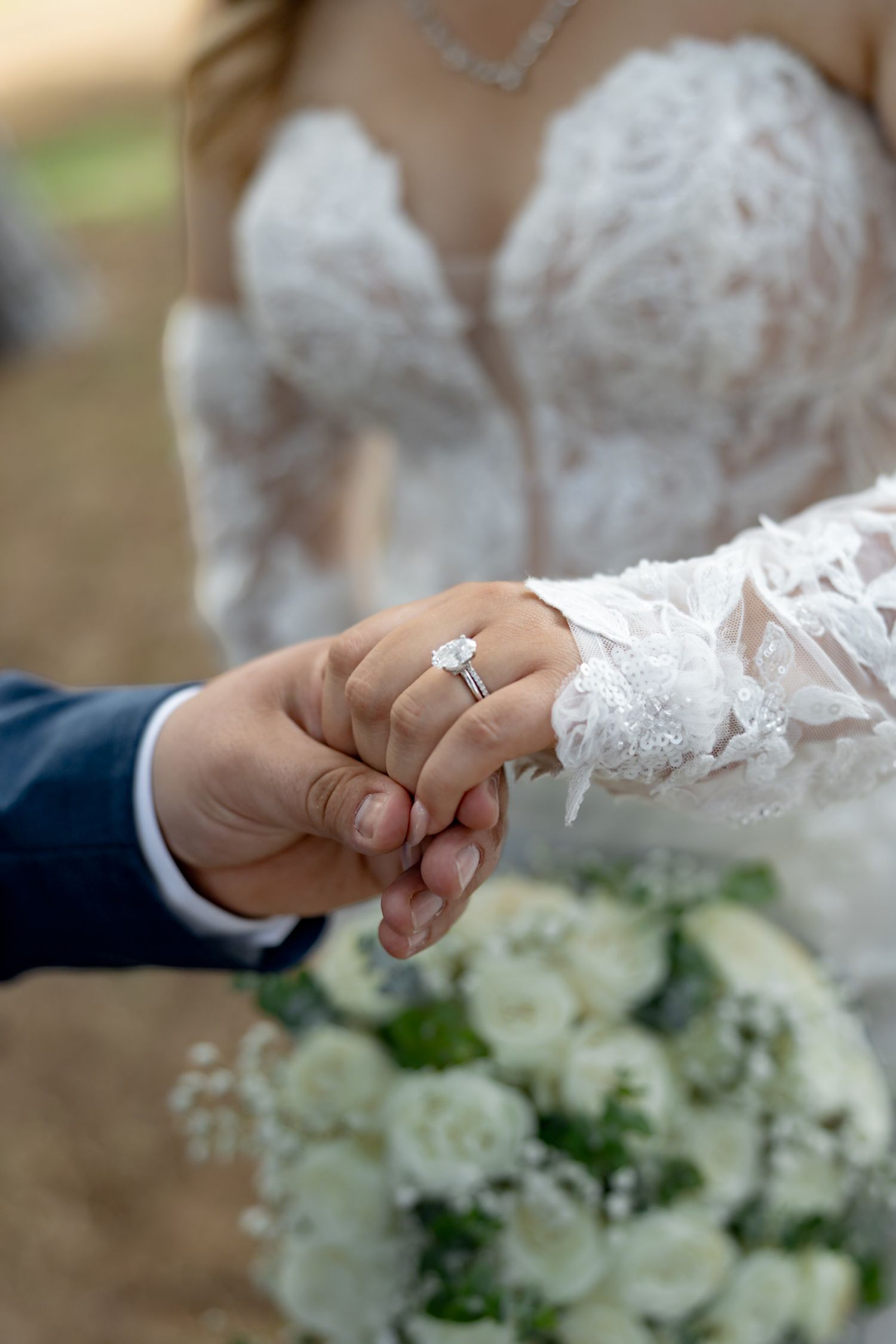 Bride and groom holding hands, showcasing a diamond engagement ring, with a white floral bouquet in the foreground.