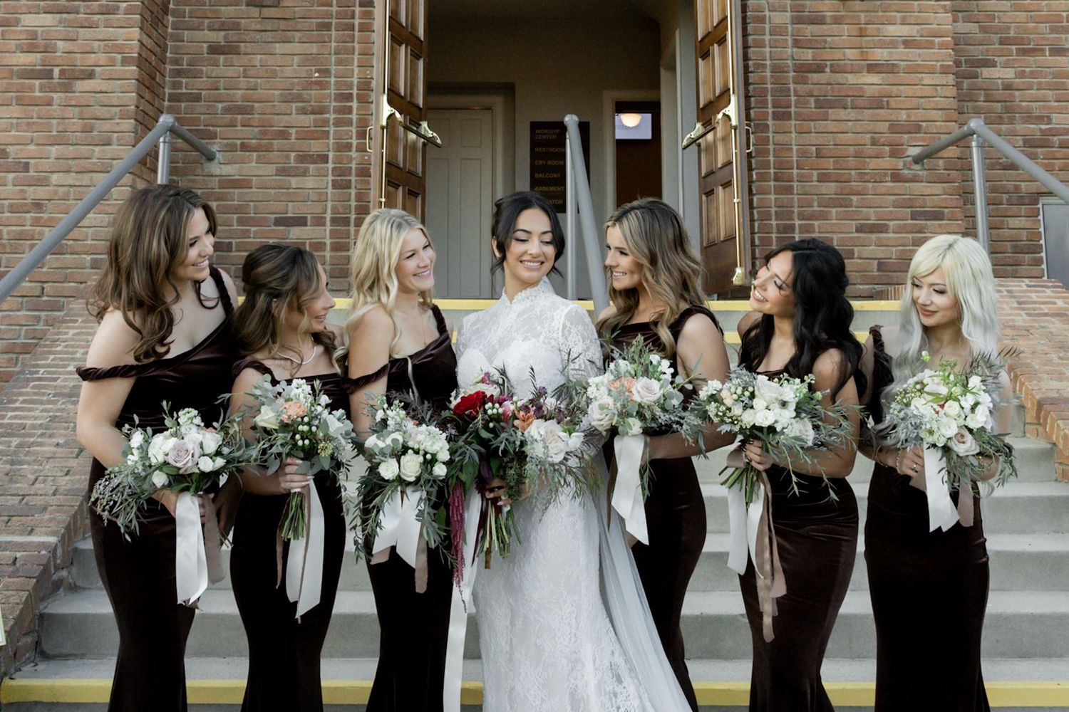 Bride in white lace gown stands with six bridesmaids in dark velvet dresses holding bouquets outside a brick church.